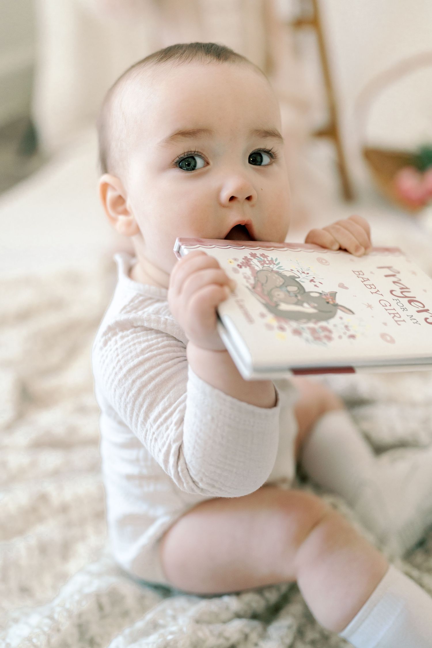 Baby in white onesie exploring a book while lying on textured bedding.