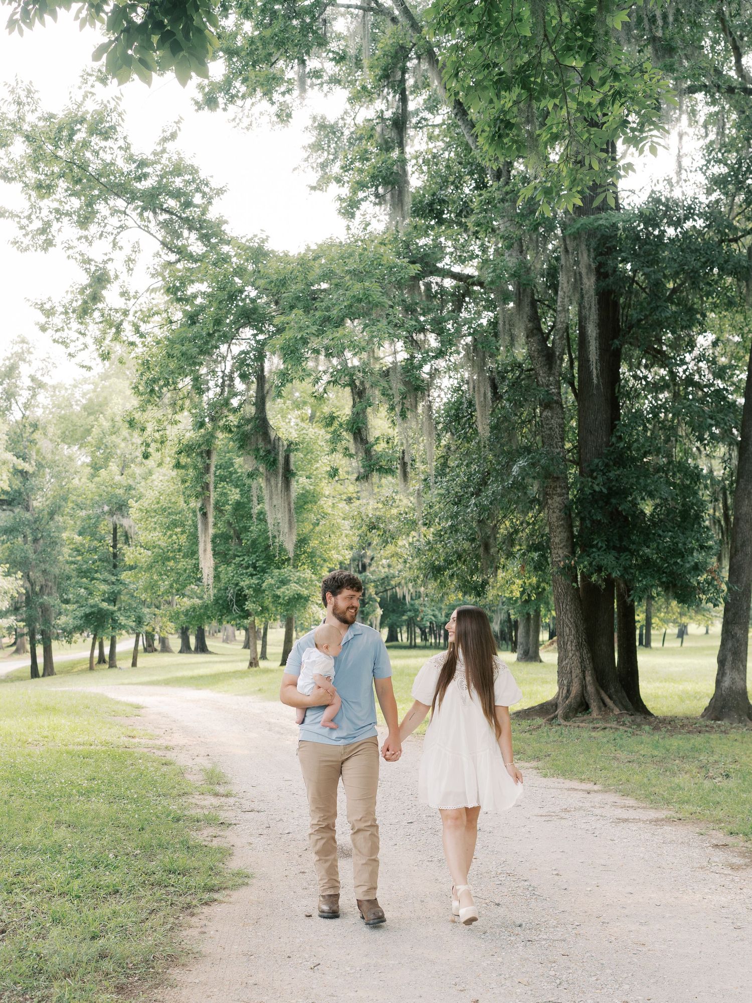 A family takes a leisurely walk along a sunlit dirt path through a wooded park.