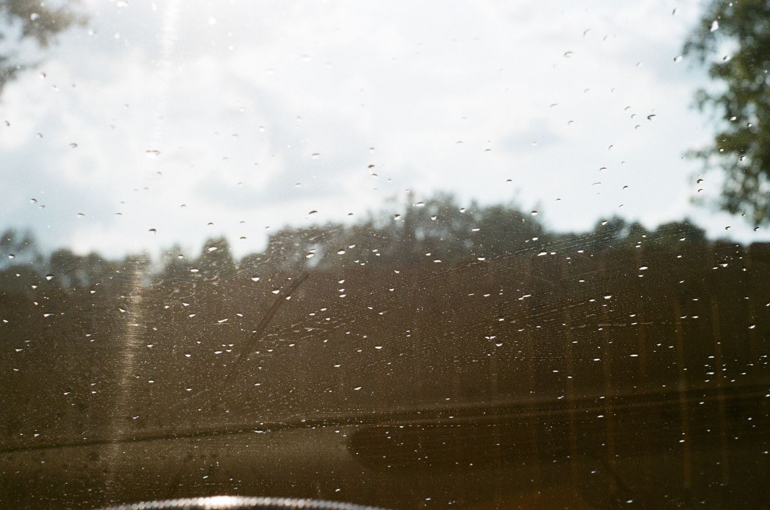 Raindrops on a car window with trees and cloudy sky visible in the background.