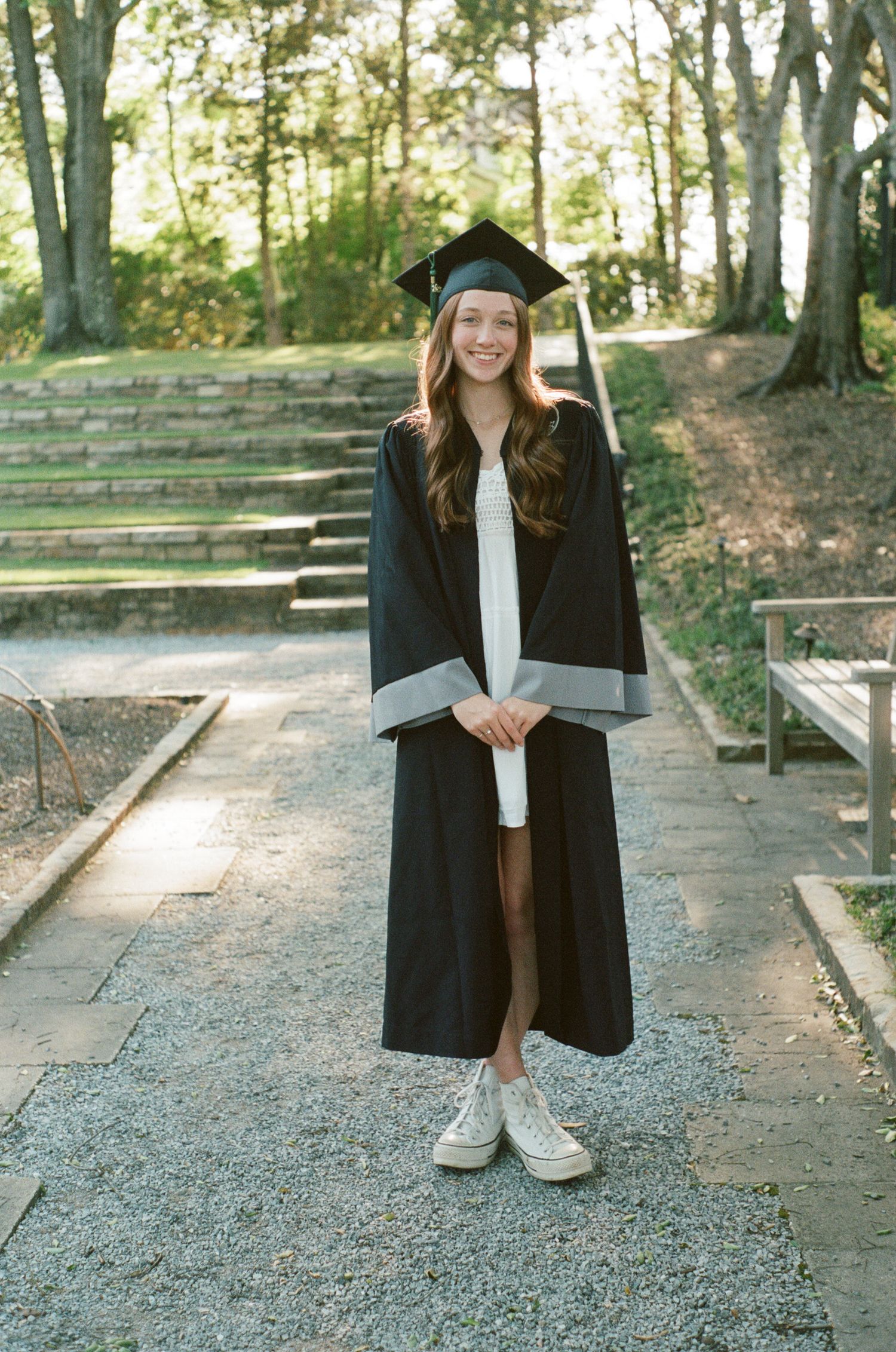 Graduate in cap and gown posing on stone steps in outdoor park setting.