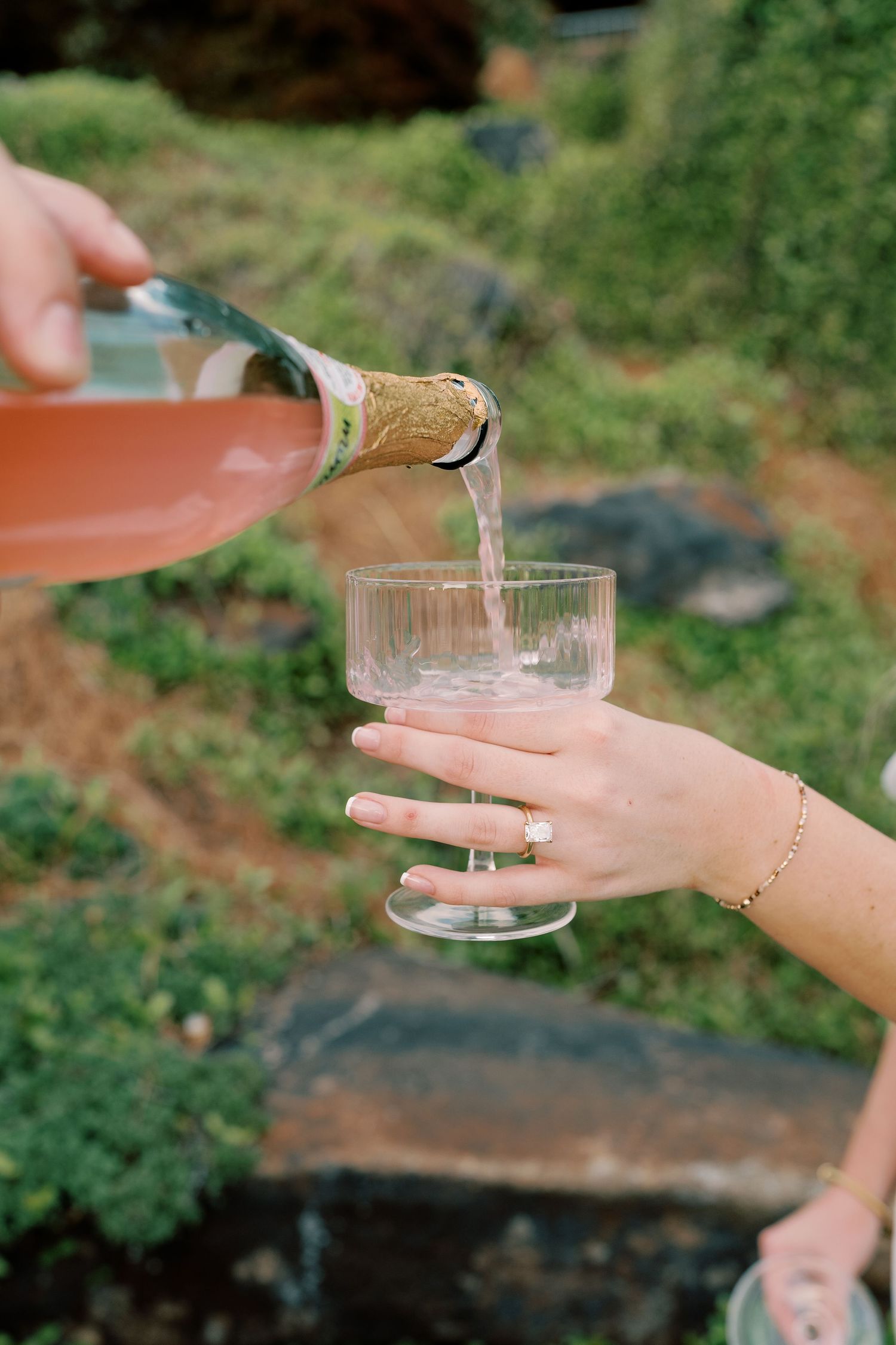 Hands pouring champagne into coupe glasses outdoors.