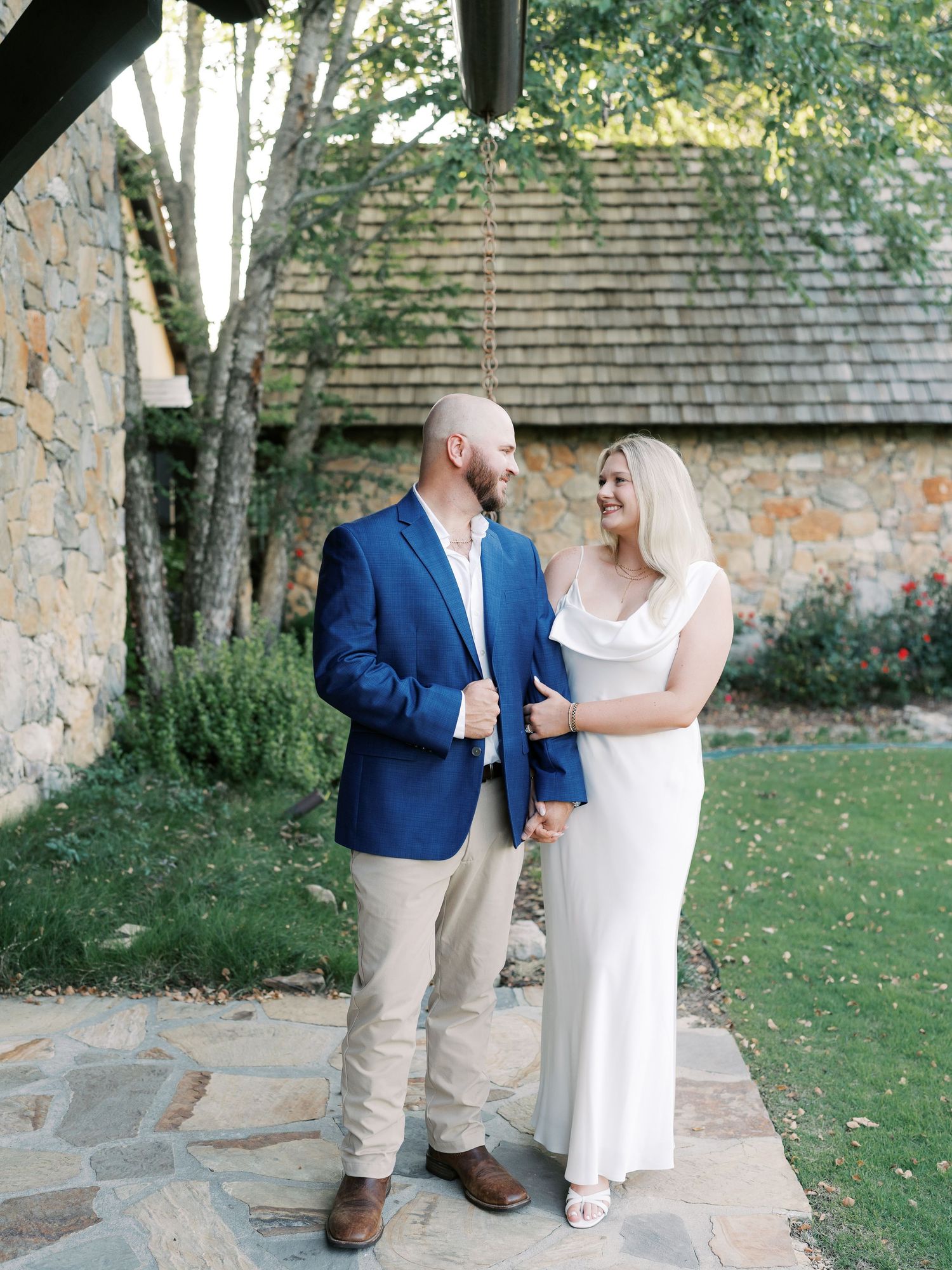 Elegant couple poses against rustic stone building in formal attire.