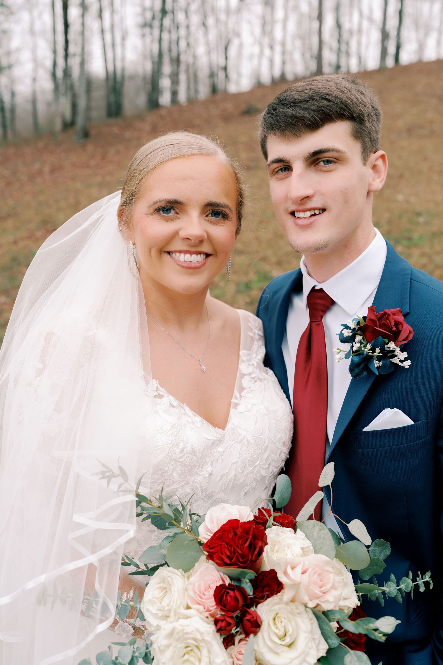 Wedding couple poses with burgundy and white floral bouquet against autumn backdrop.
