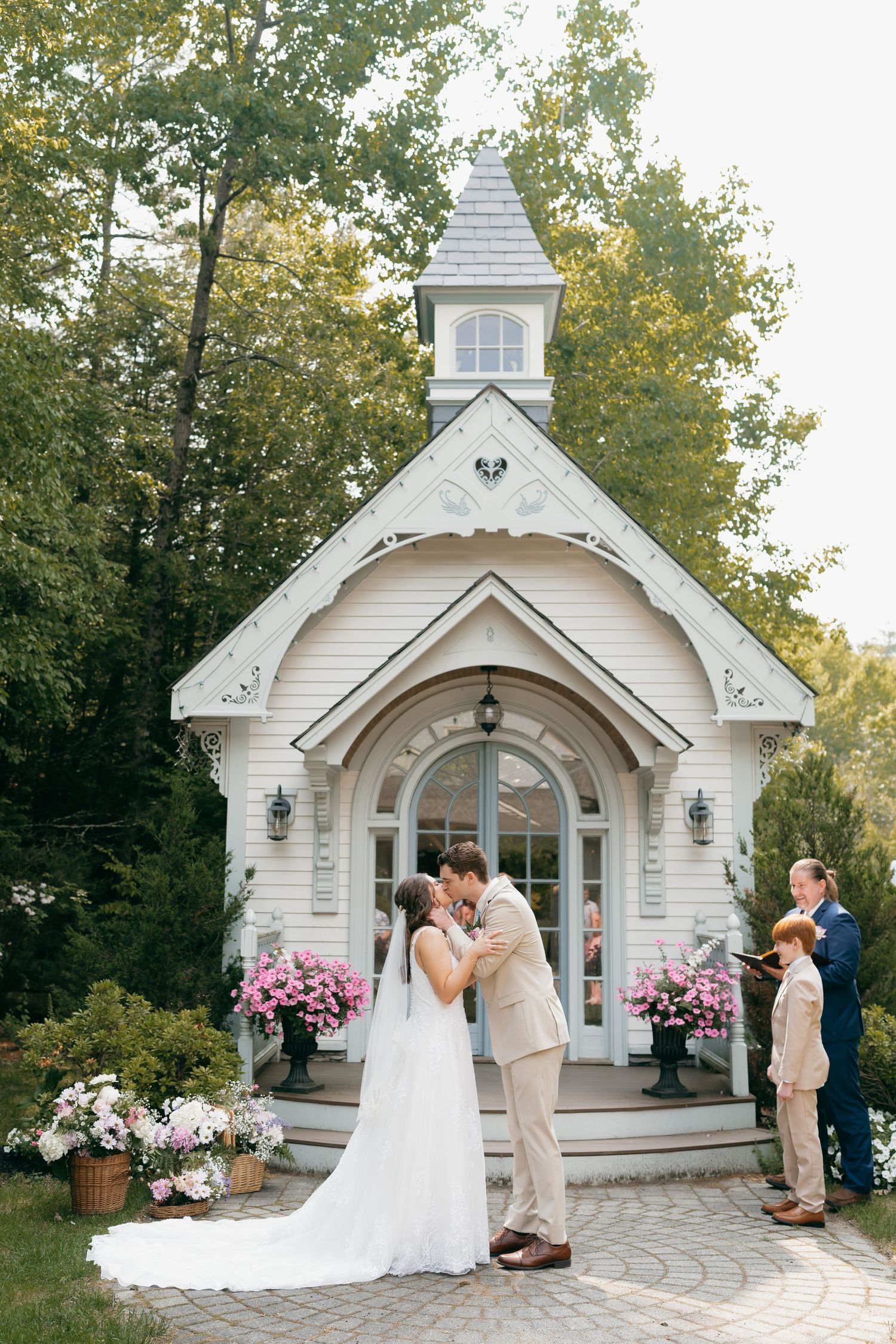 A charming white chapel with decorative trim and pink flower arrangements frames a wedding ceremony.
