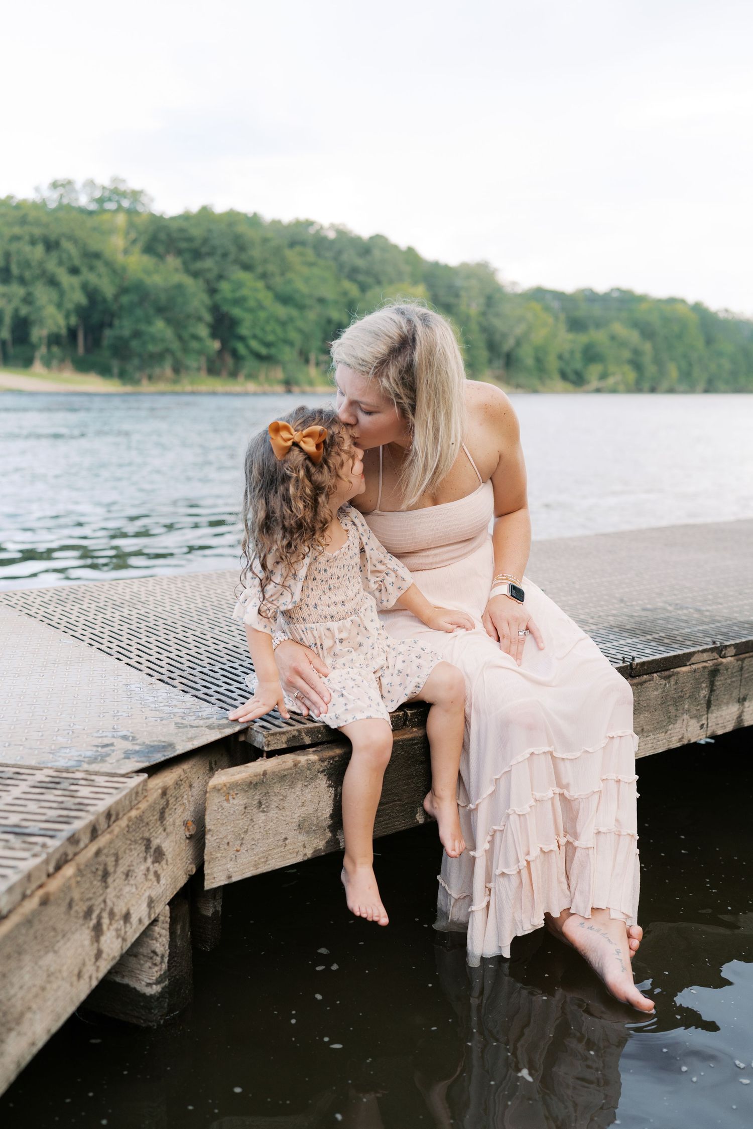 Two people in light-colored dresses sit together at the edge of a wooden dock over water.