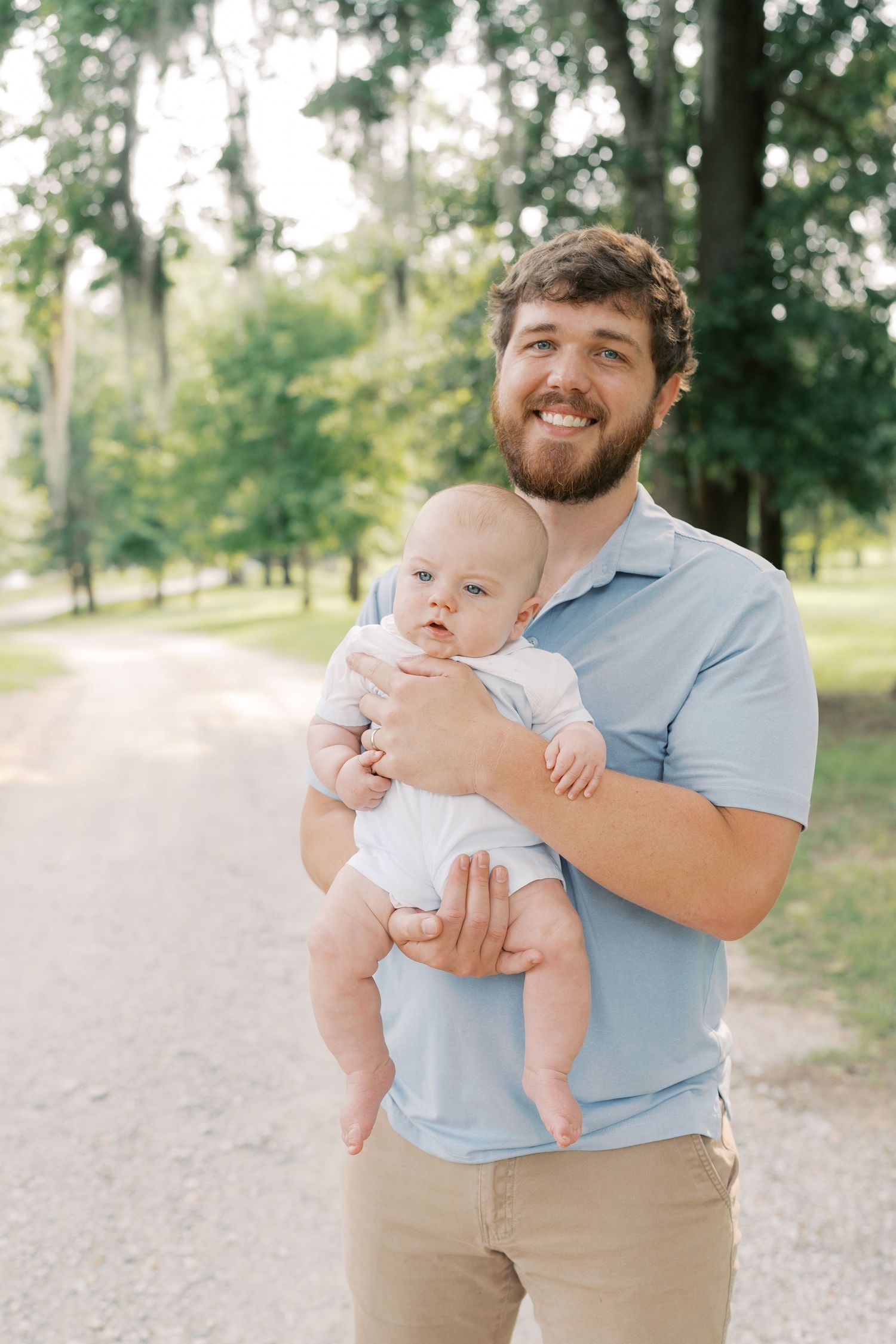 A parent holds a small baby while standing on a tree-lined path.