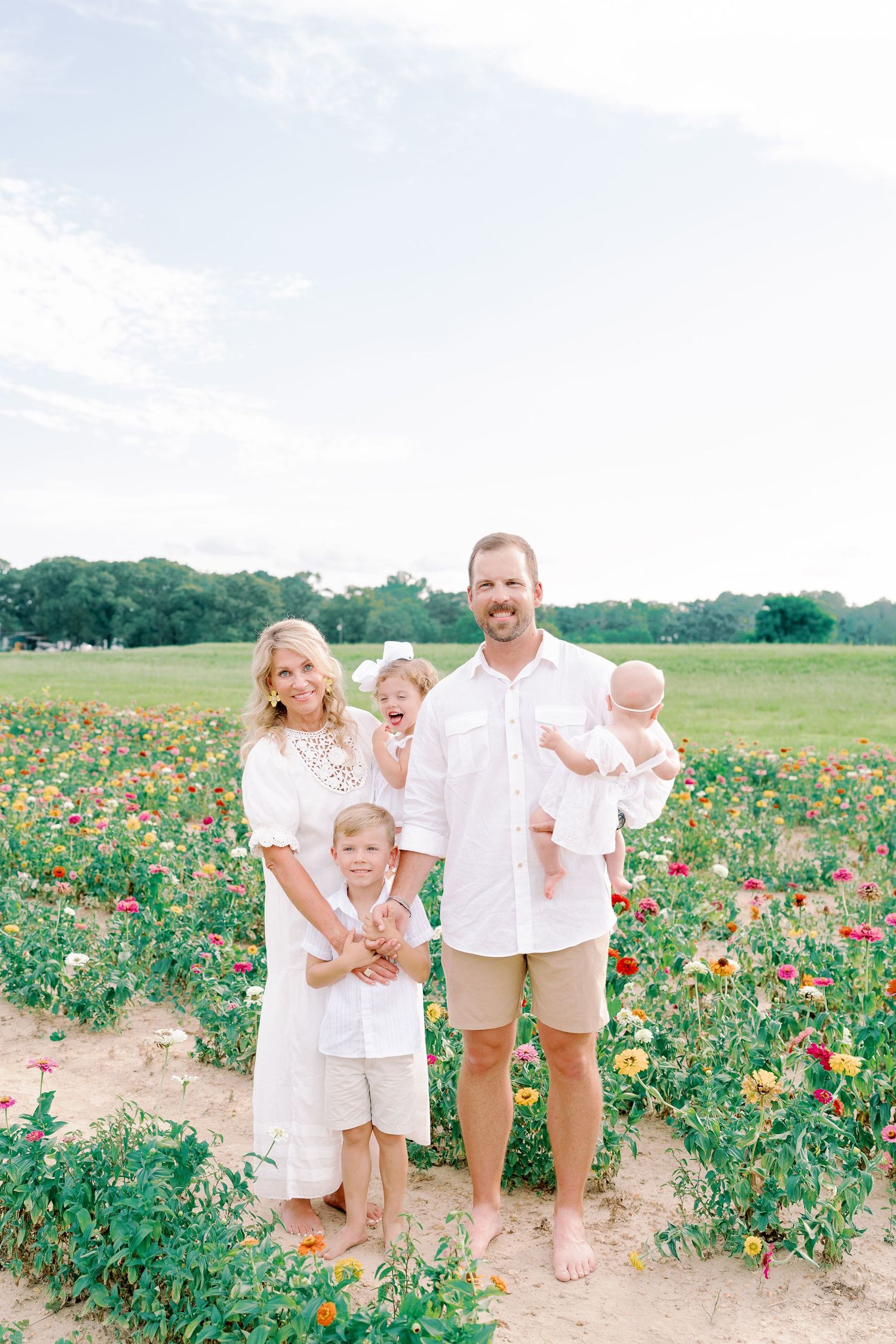 Family wearing coordinated white outfits poses in field of vibrant summer flowers.