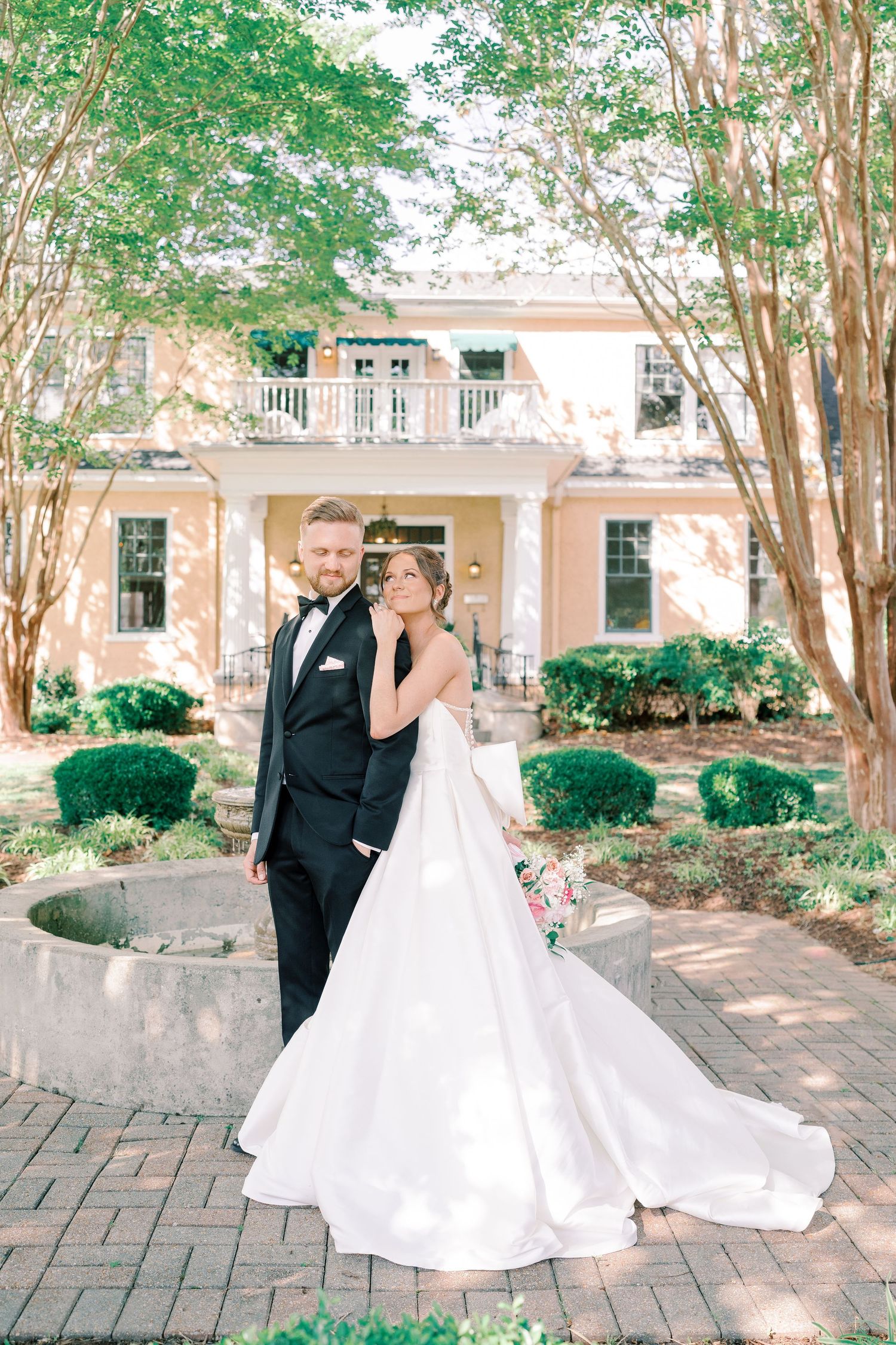 Wedding couple twirling on brick courtyard surrounded by manicured trees and colonial architecture.