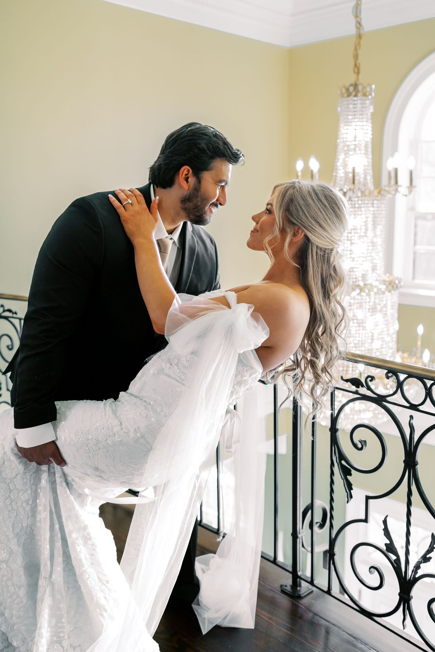 Romantic wedding couple poses in elegant interior with ornate railings and chandelier.