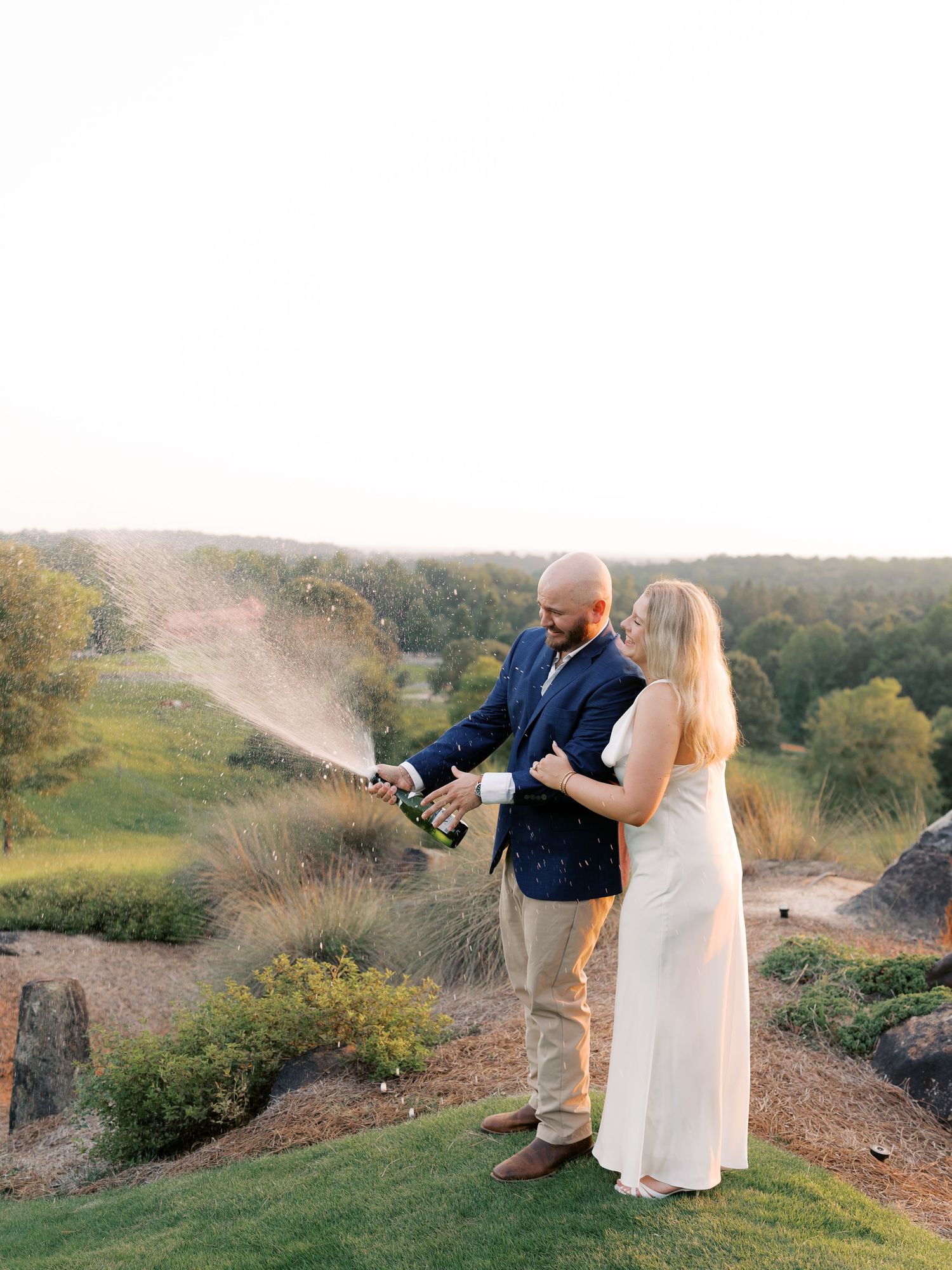 A couple pops champagne on a scenic hillside overlooking a green landscape at sunset.