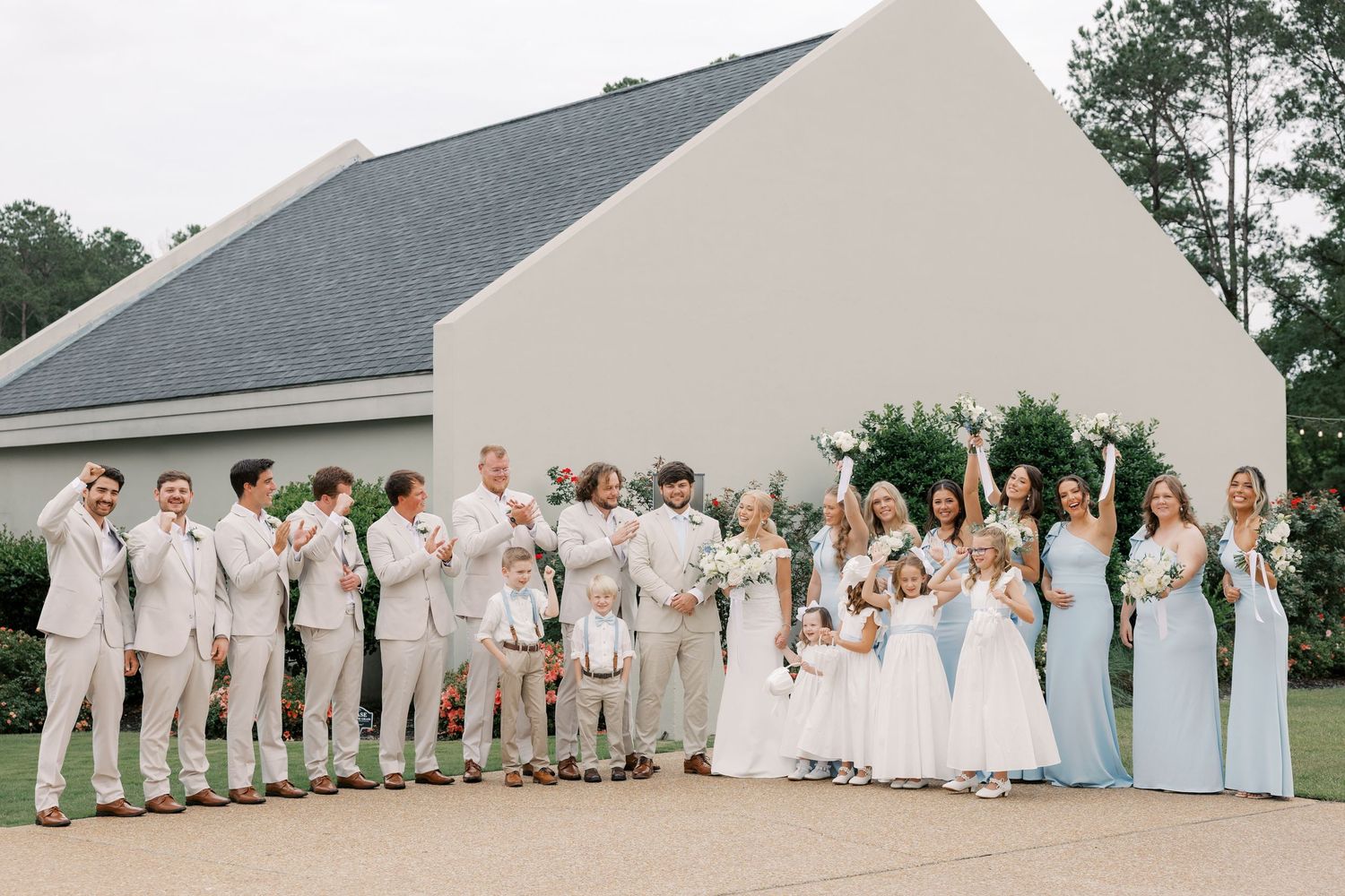 Wedding party lined up outdoors against white building in light blue and cream attire.