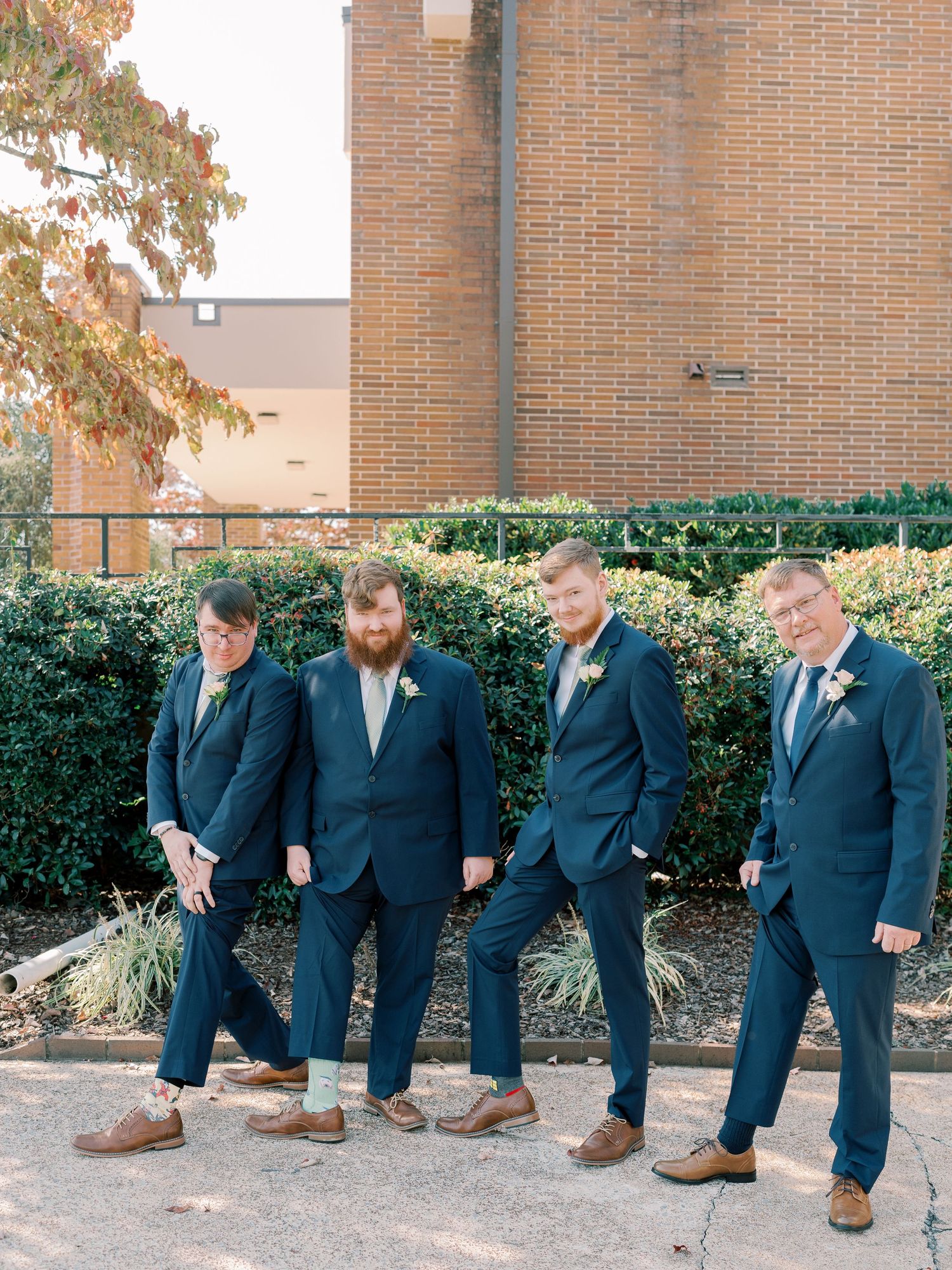 Groomsmen in navy suits posing casually against brick wall outdoors.