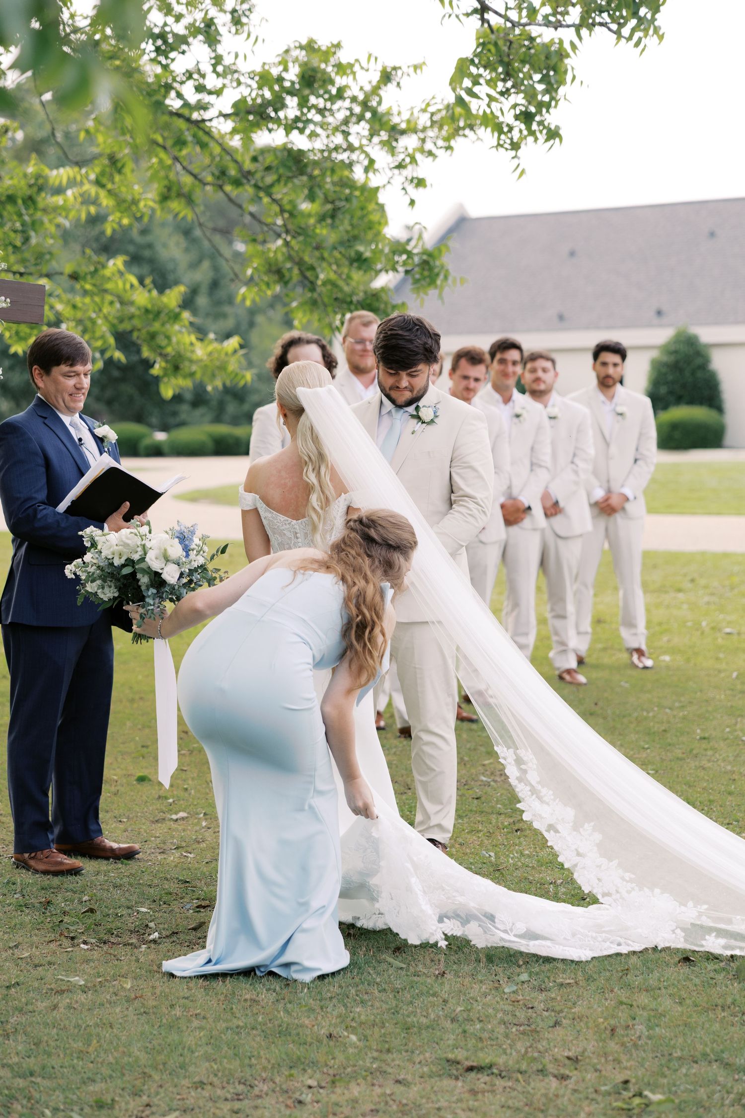 Wedding party members in white and blue attire pose on a grassy lawn.