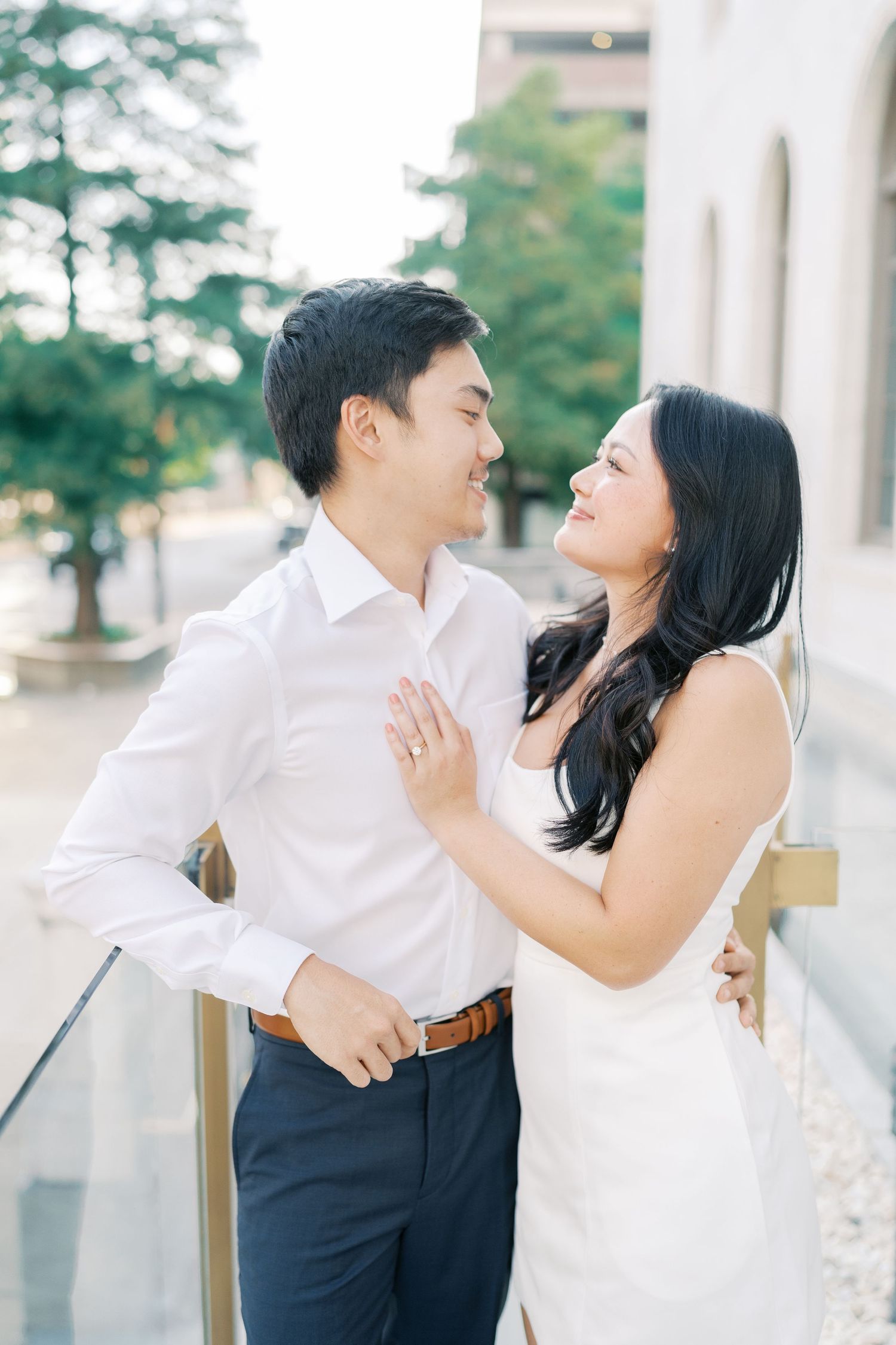 A romantic couple embraces against a light-colored building exterior with columns and greenery in the background.