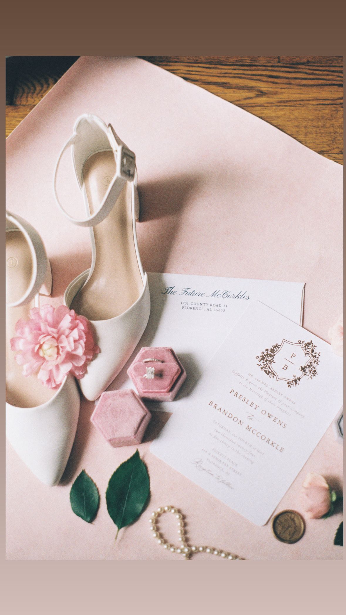 Wedding invitation suite with pink accessories and white shoes on wooden surface.