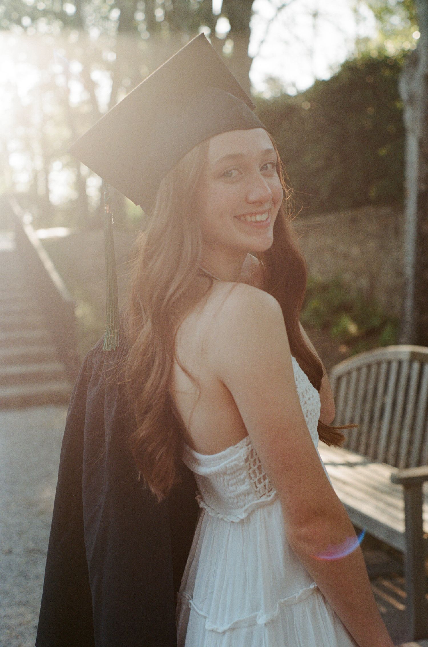 Sunlit graduation portraits in white dress and black cap against natural backdrop.