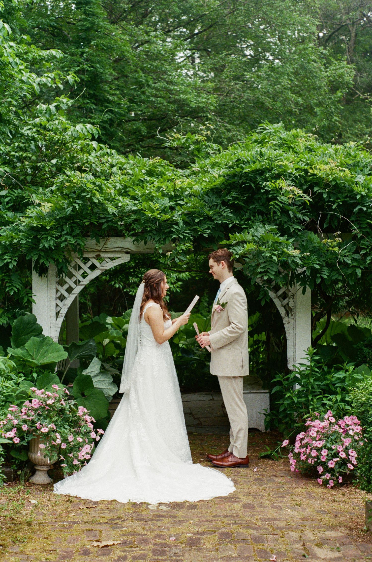 A couple exchanges vows under a vine-covered garden arch decorated with pink flowers.