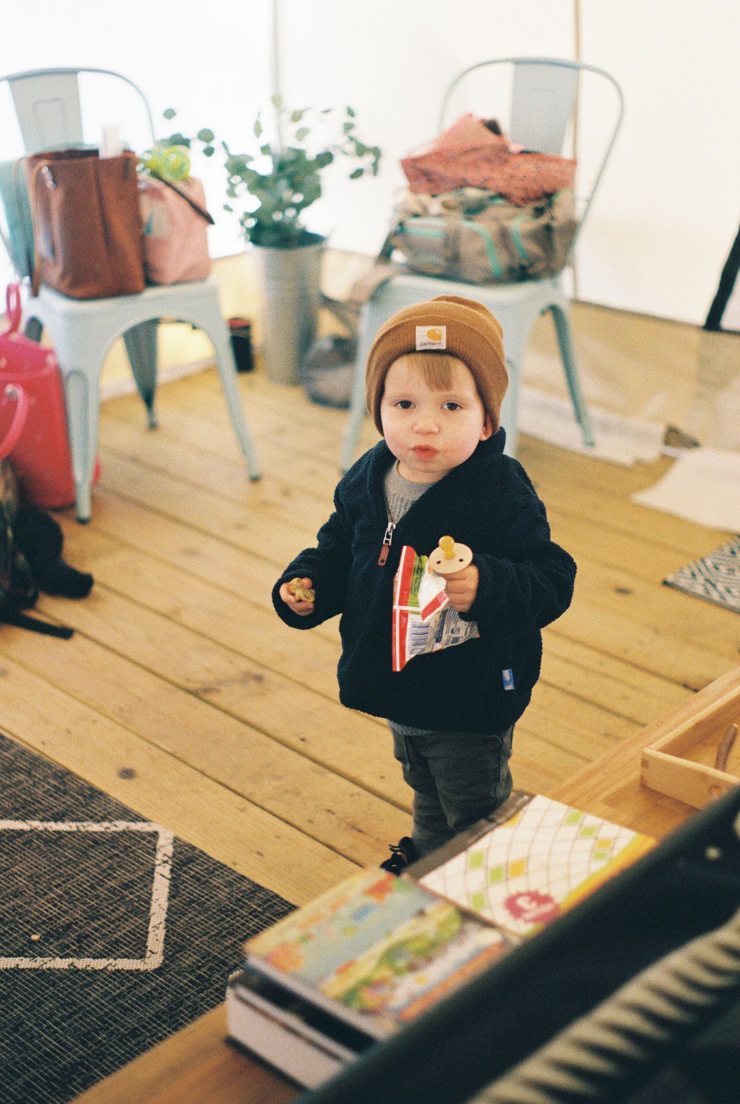 Child in brown beanie and black jacket exploring books on wooden floor with potted plants in background.