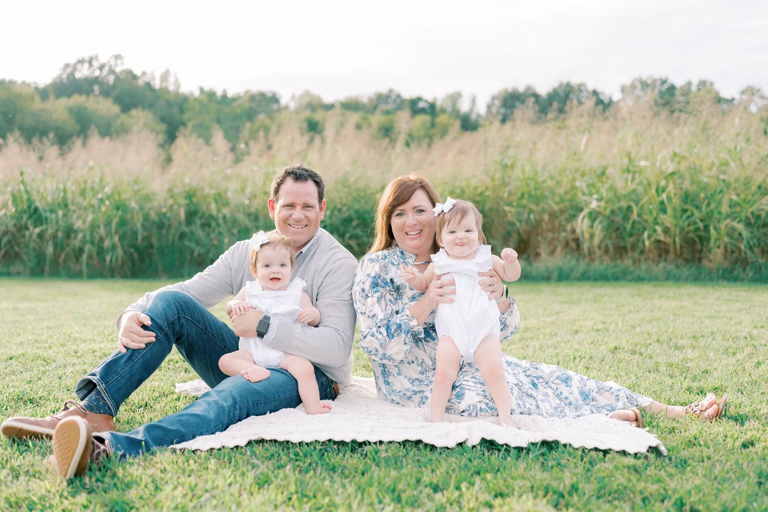 Family relaxing together on a white blanket in a sunny meadow with tall grass in the background.