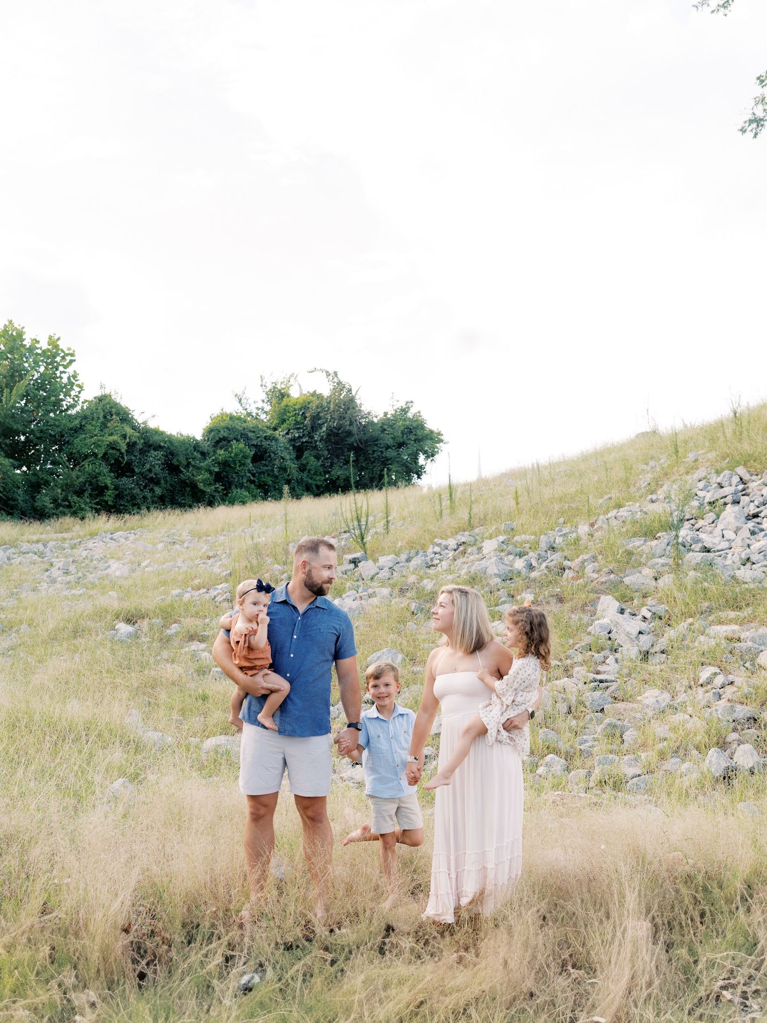 A family group poses candidly in a summer field with tall grass and rocks.