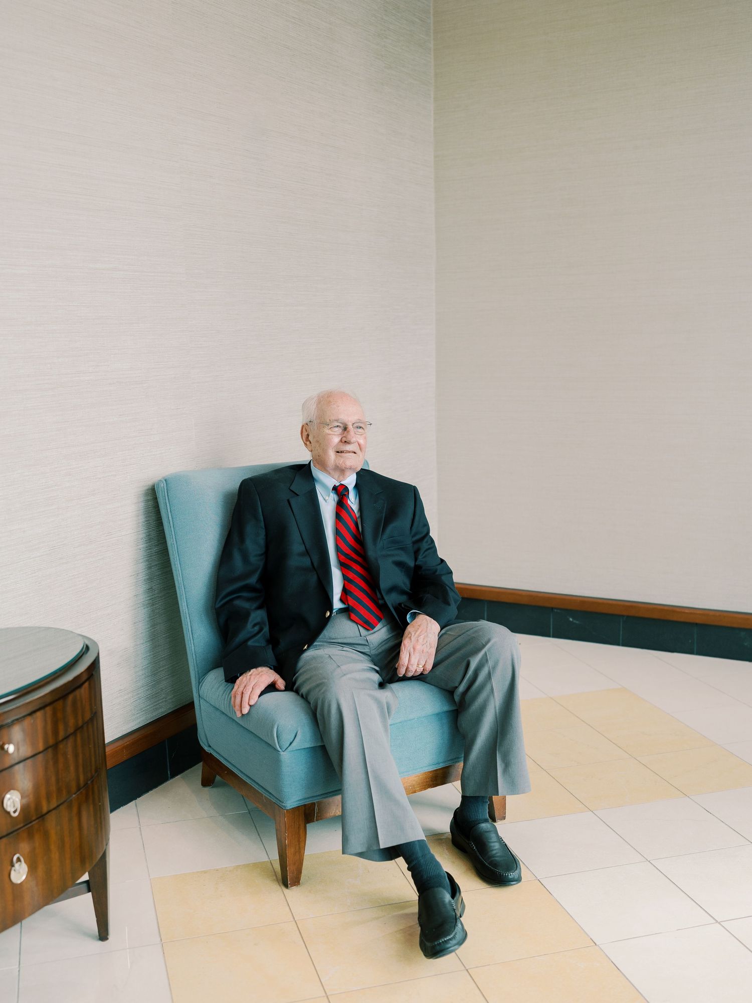 Senior in dark suit sitting on light blue chair against white wall.