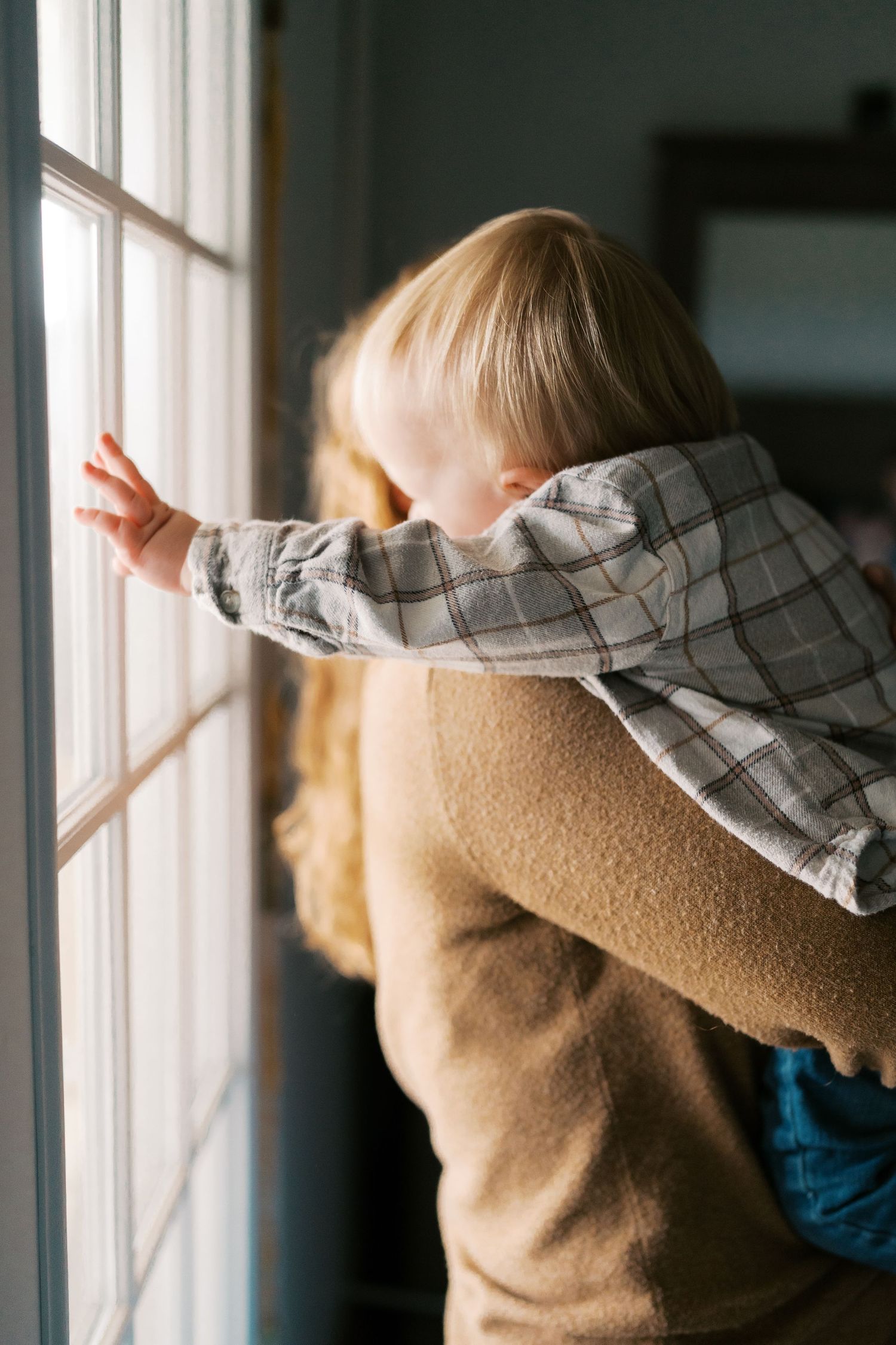 Child looking out sunny window while wearing plaid shirt and tan sweater.
