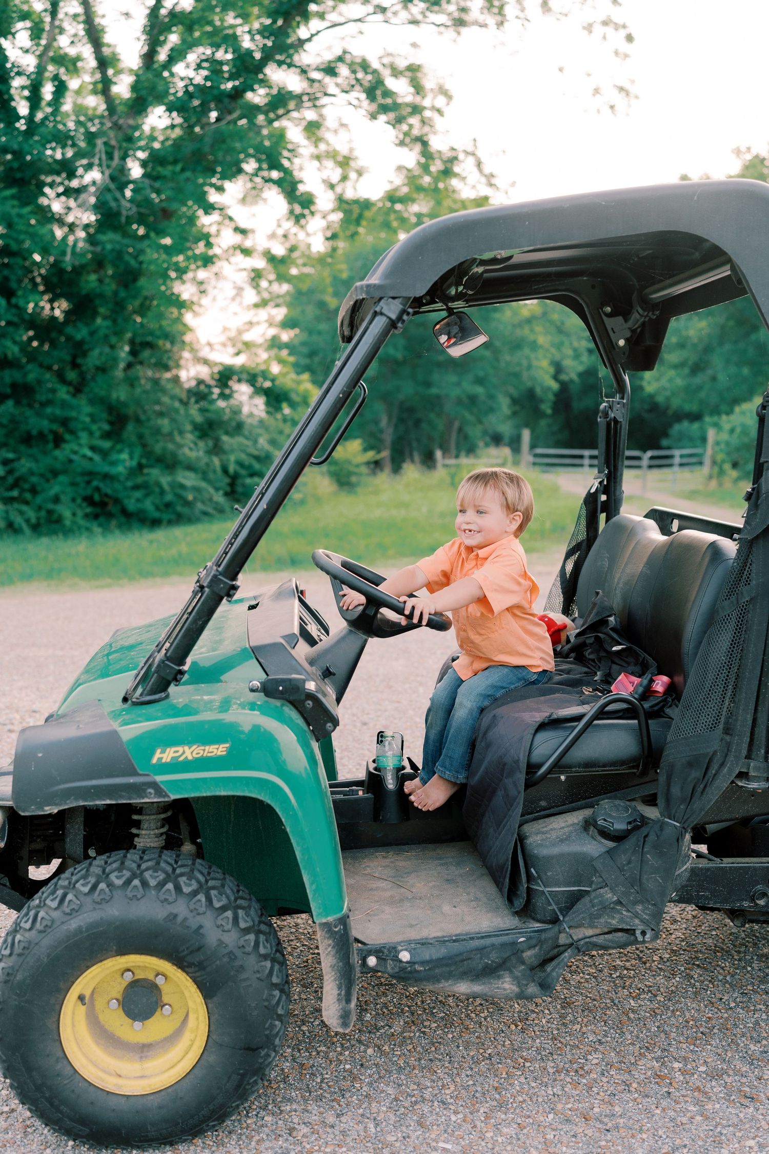 Child sitting in green John Deere utility vehicle on gravel path.
