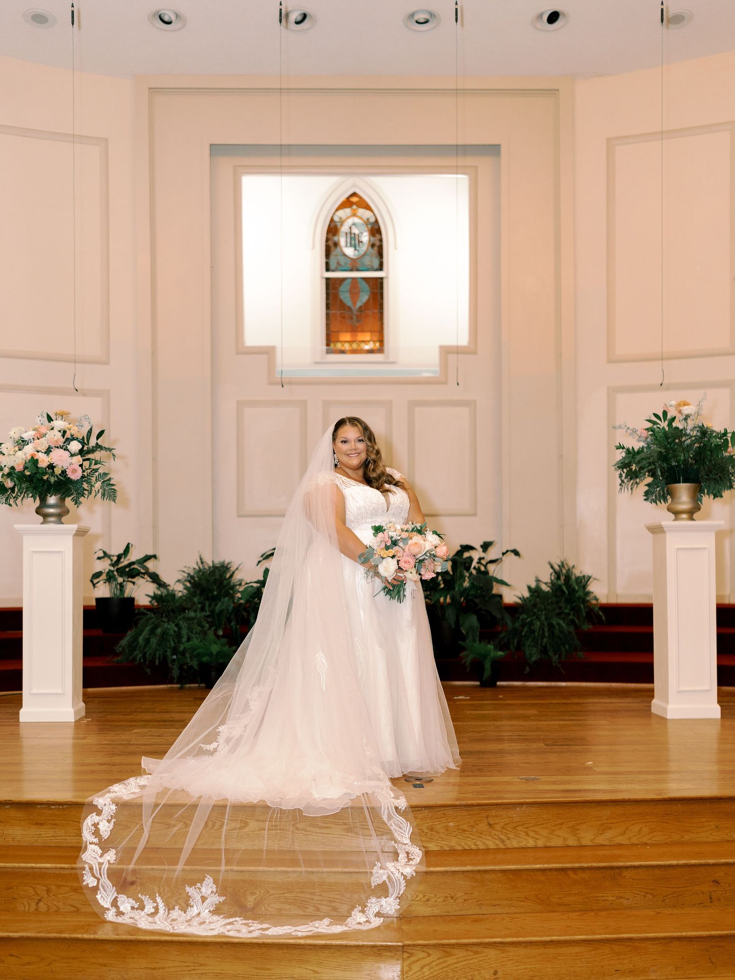A wedding ceremony setup in a chapel with white columns, floral arrangements, and a beautiful long veil trailing on hardwood floors.