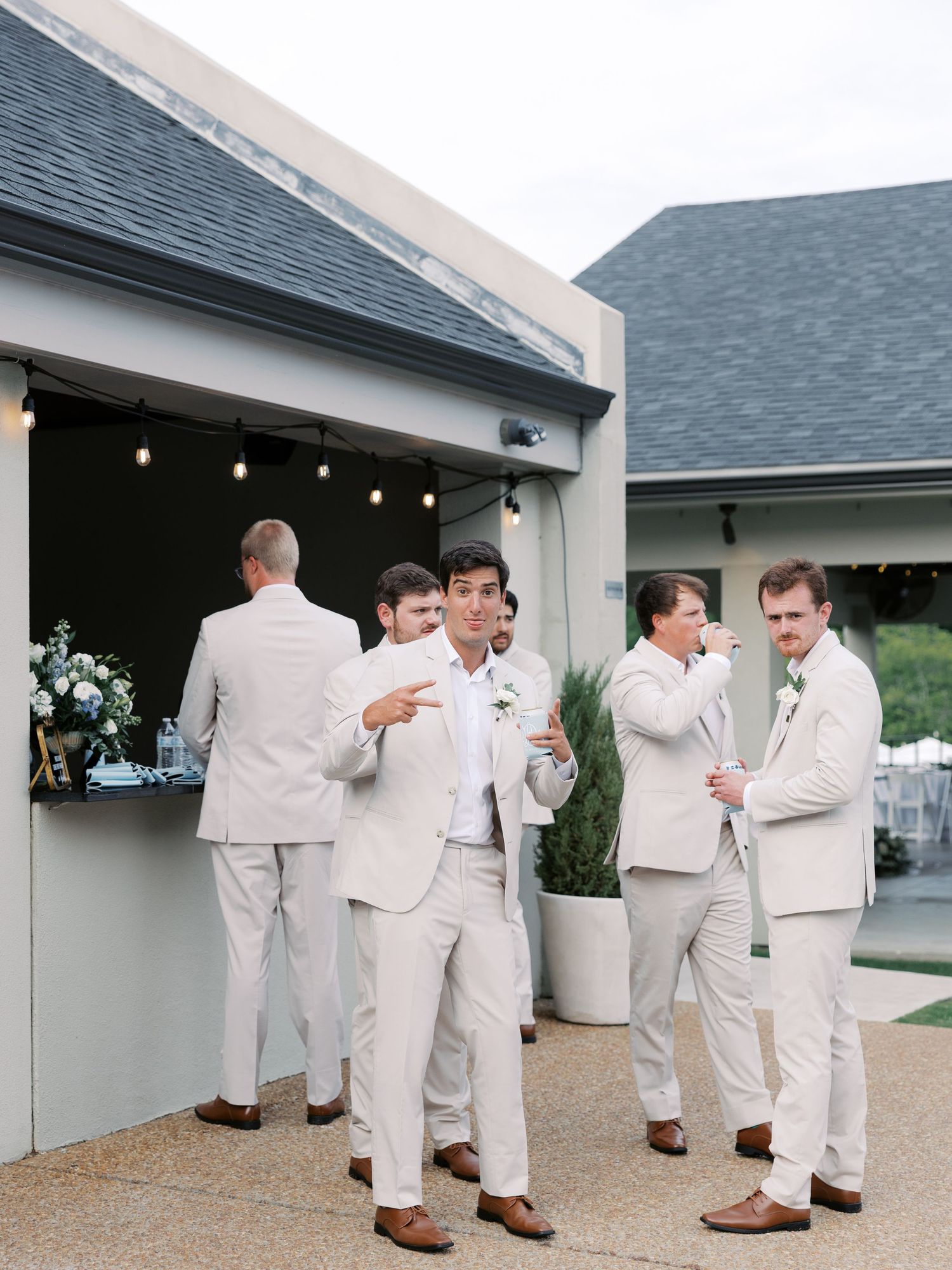 Groomsmen in light beige suits gathered outside venue with string lights.
