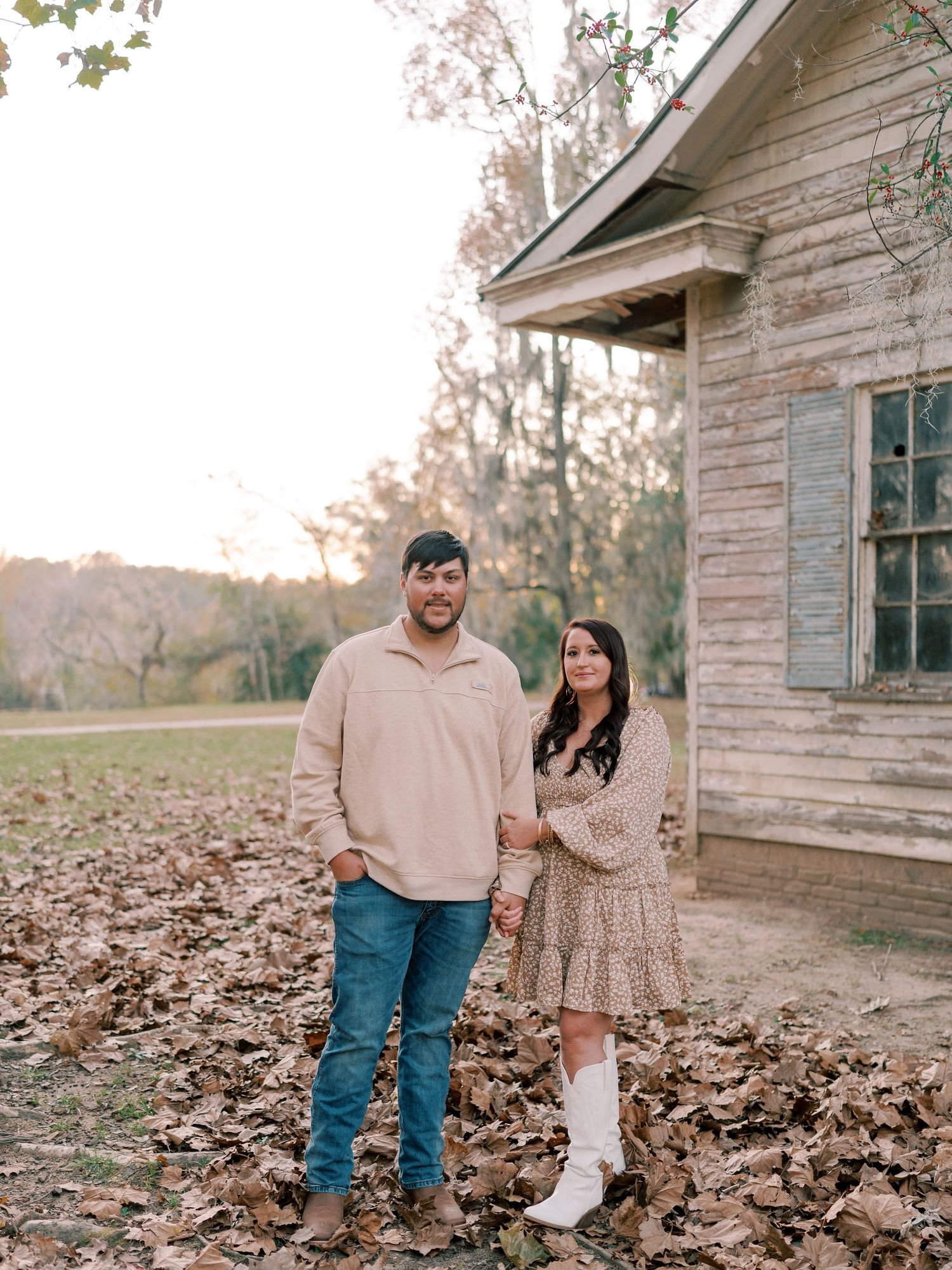A couple stands together on fallen leaves near a weathered wooden building.