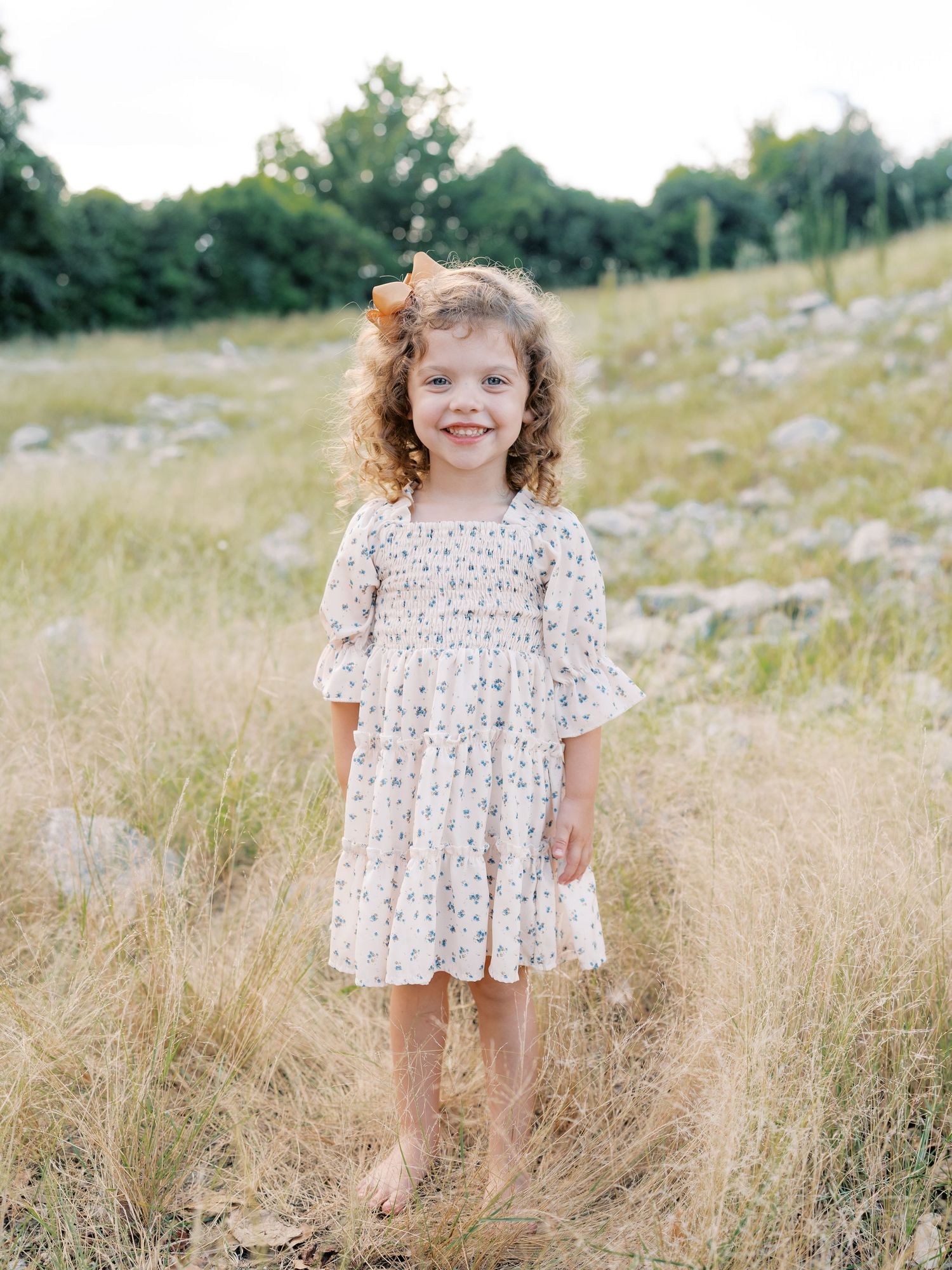 Young child in a floral dress standing barefoot in a summer meadow.