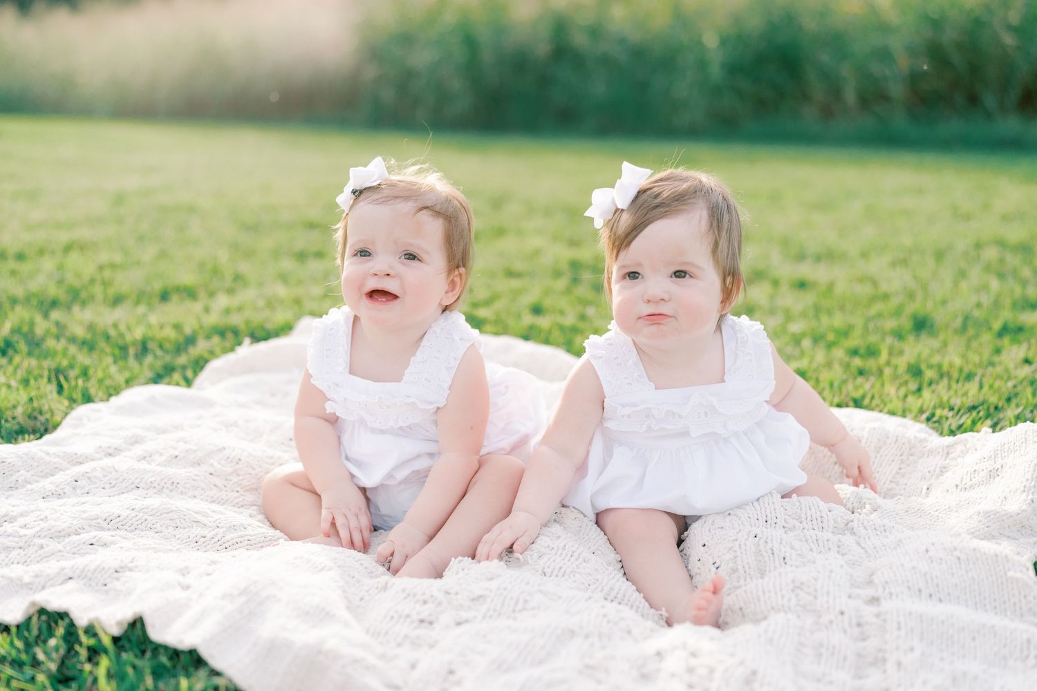 Two babies sitting together on white blanket in grassy field during sunset.