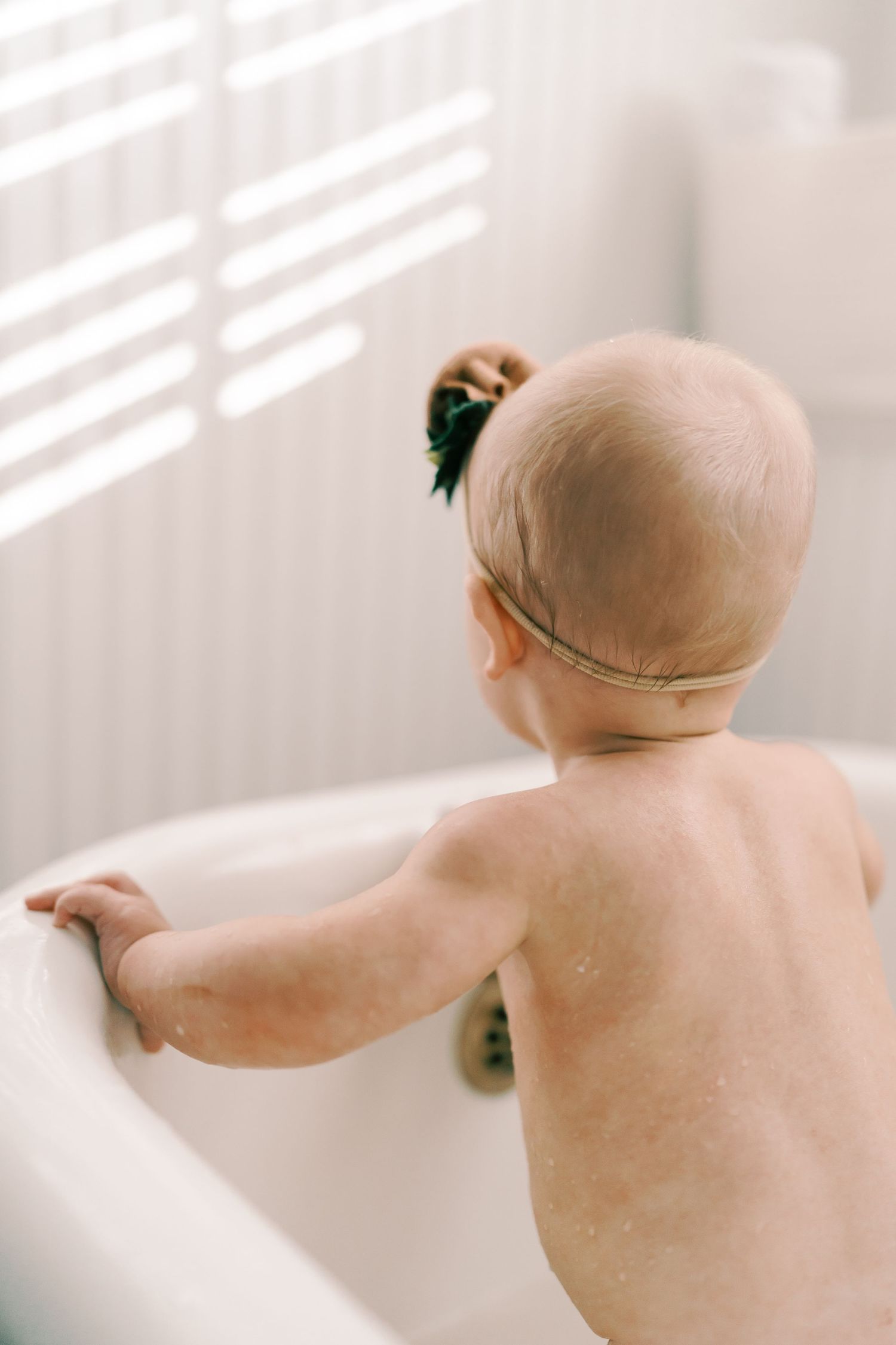Baby reaches out while playing in sunlit white bathroom.