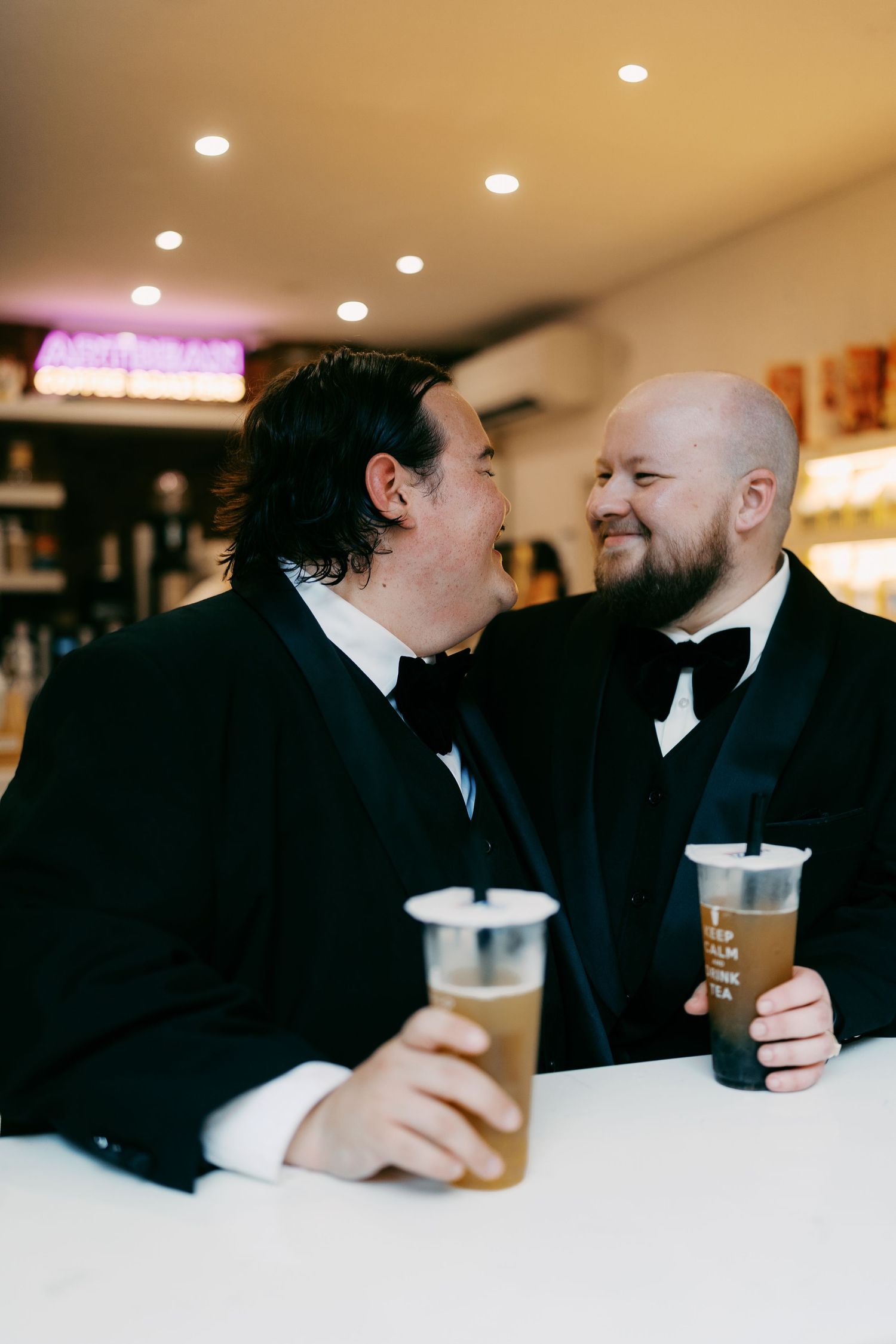 Two people in black suits share drinks at a bar counter with overhead lighting.