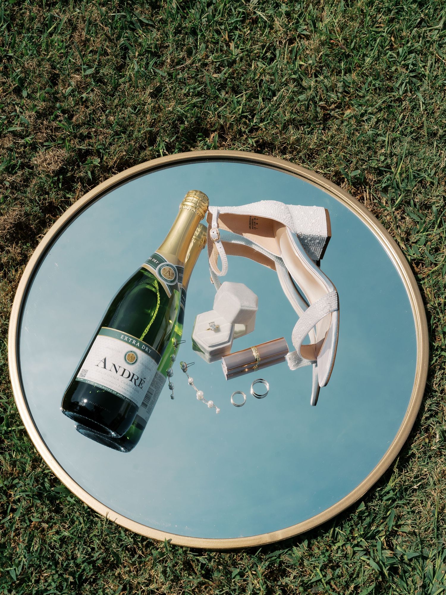 Wedding details styled on an oval mirror with champagne bottles and rings against grass background.