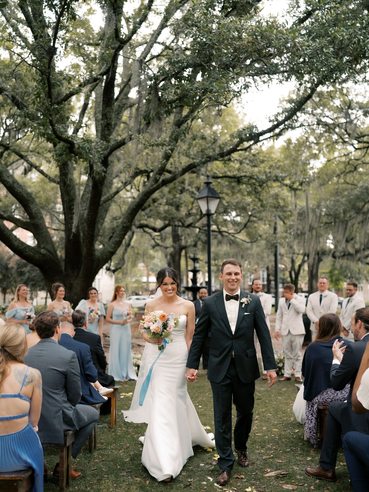 Wedding guests gather under large oak trees during an outdoor ceremony.