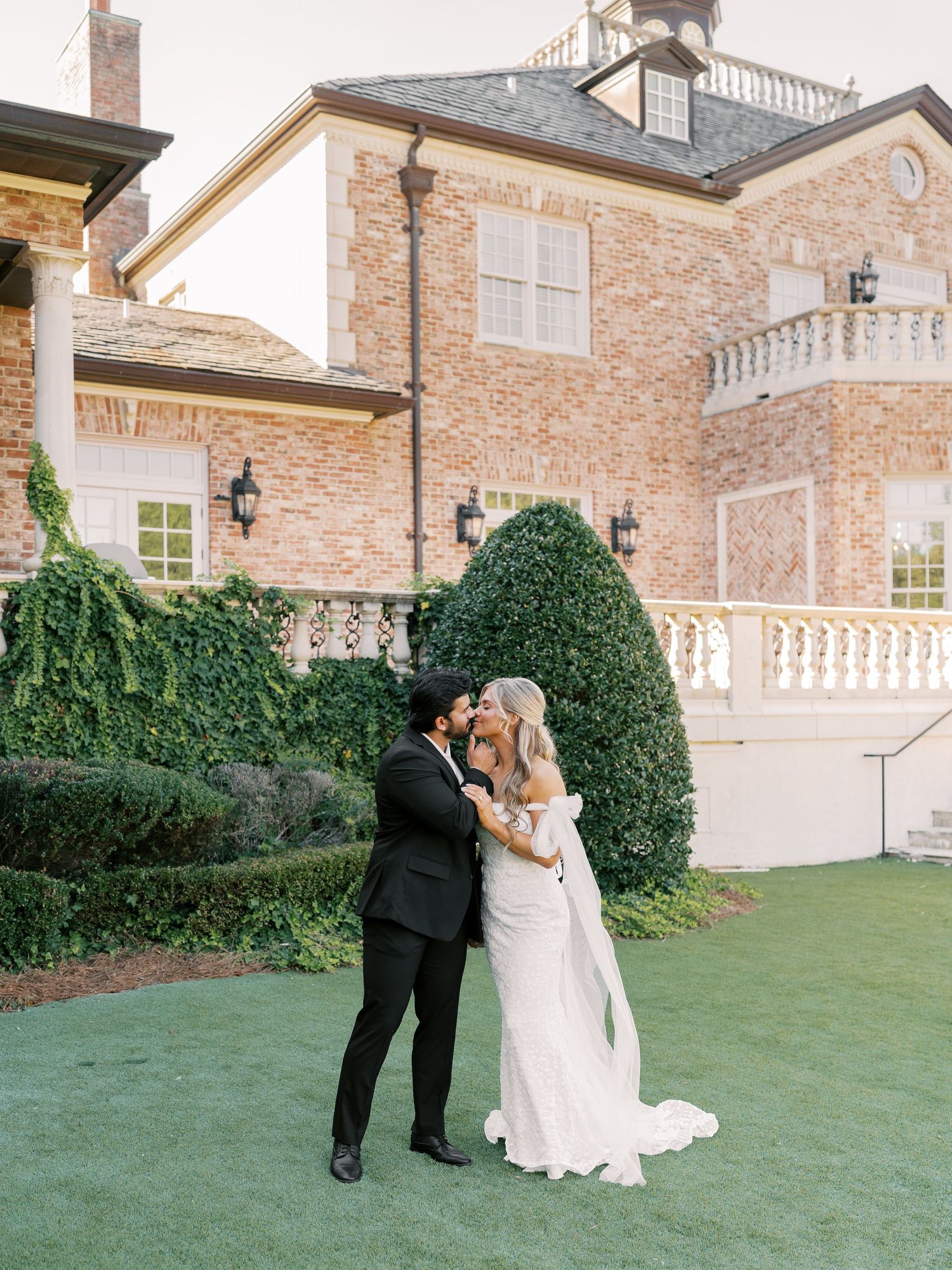 A romantic moment captured outside a grand brick estate with manicured grounds.
