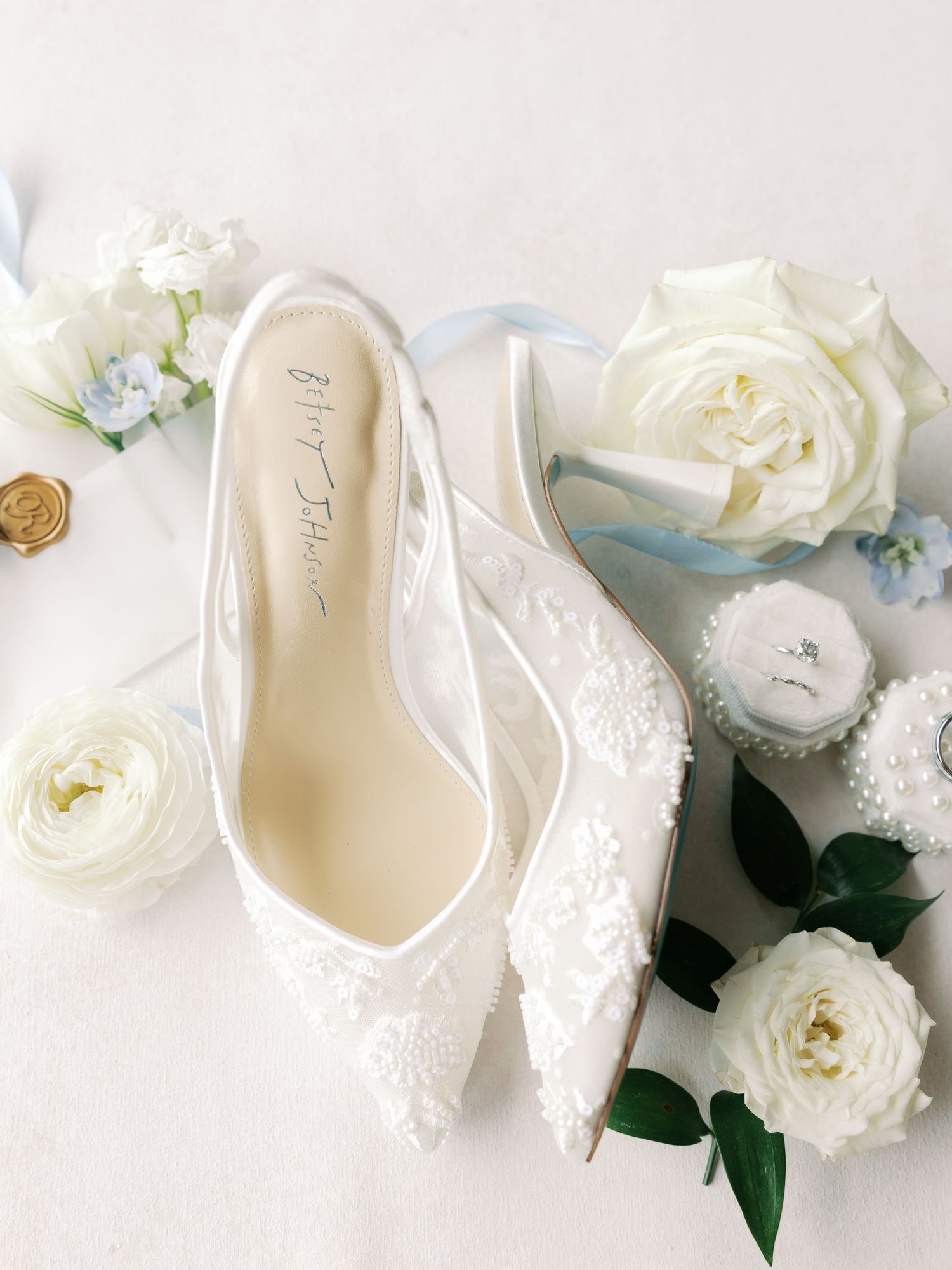 Elegant white bridal shoes surrounded by delicate white roses and greenery on light background.