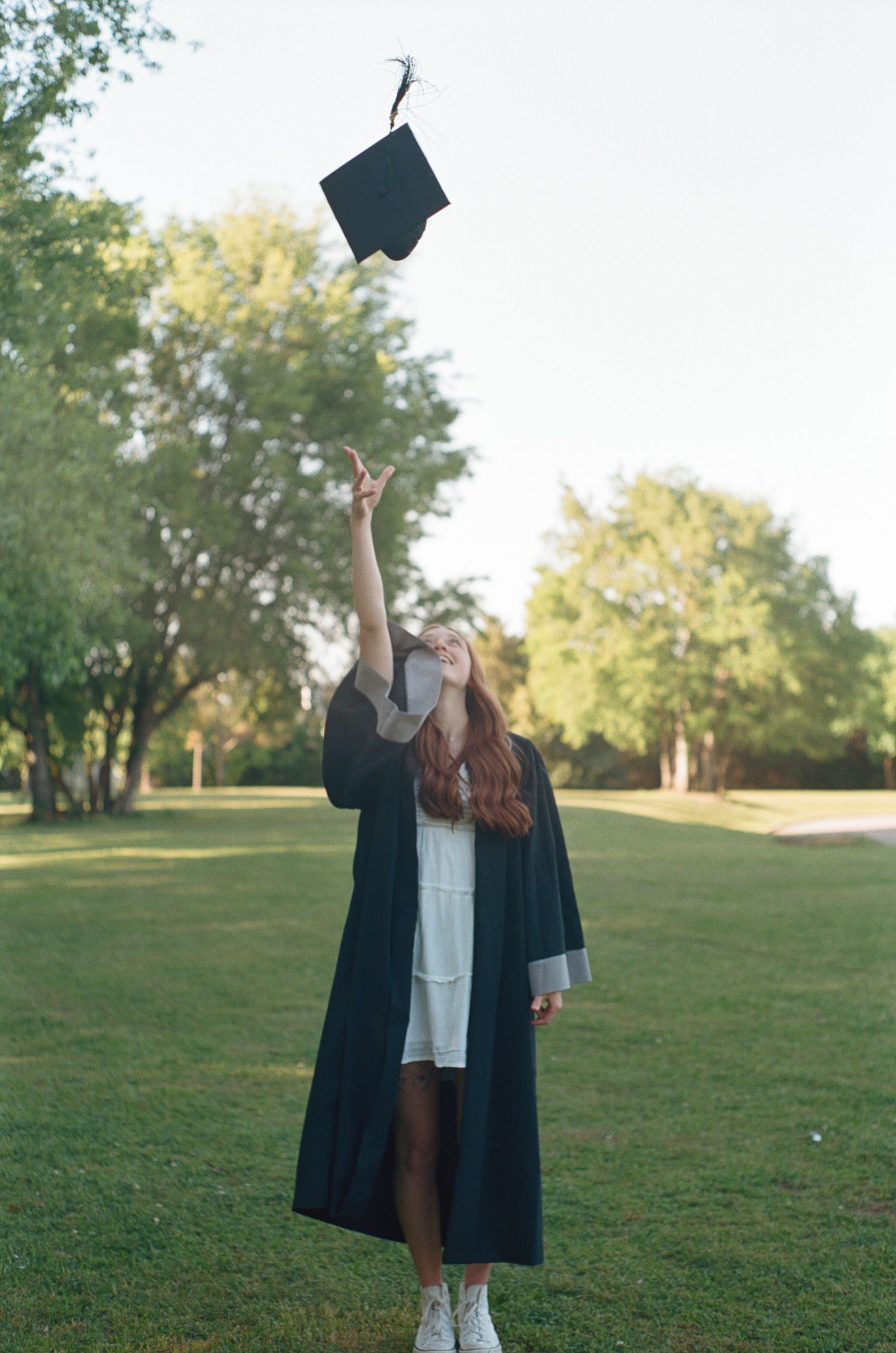 Graduate tossing cap in air on grassy hill during celebration photoshoot.