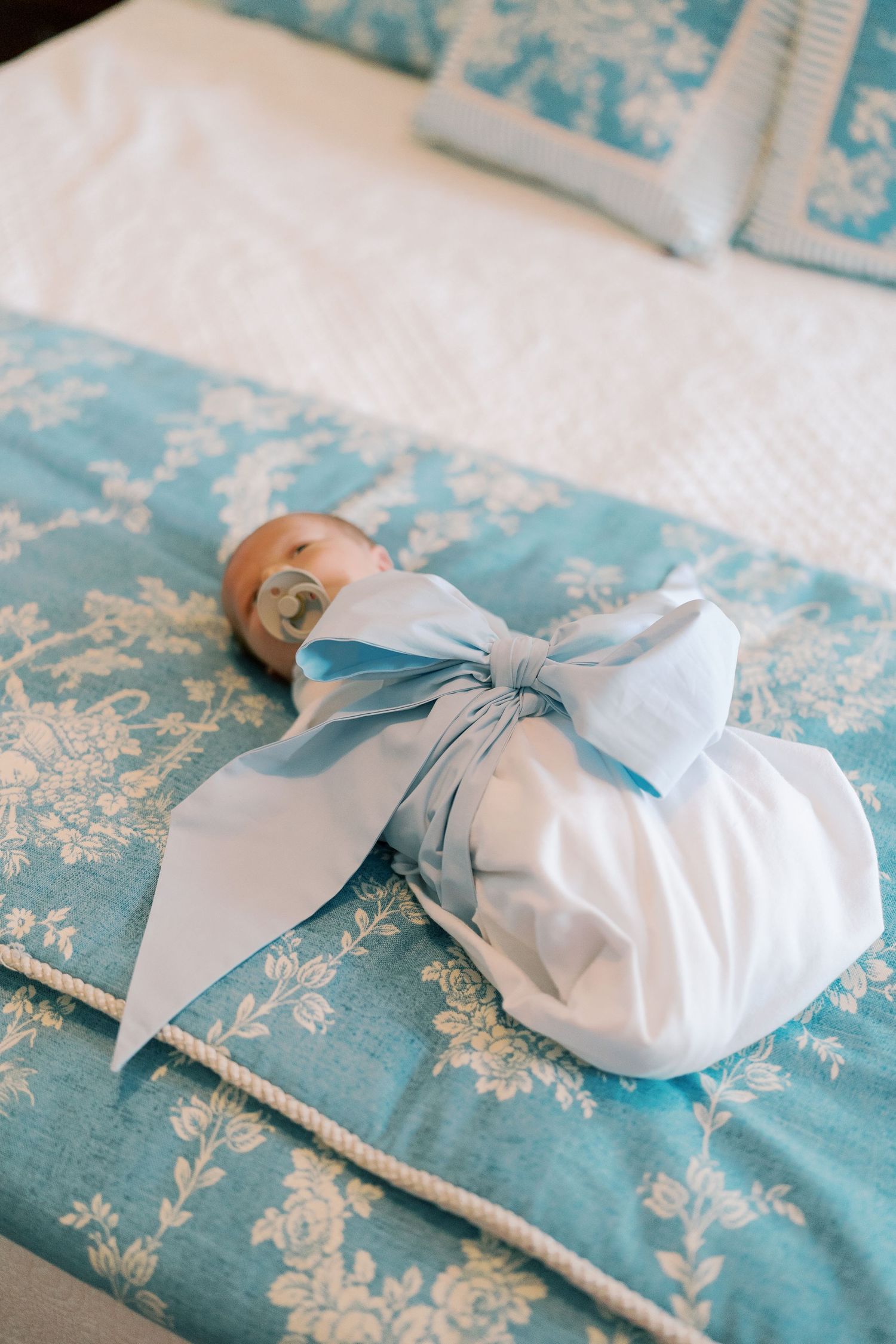 Baby swaddled in light blue bow rests on floral patterned bedding.