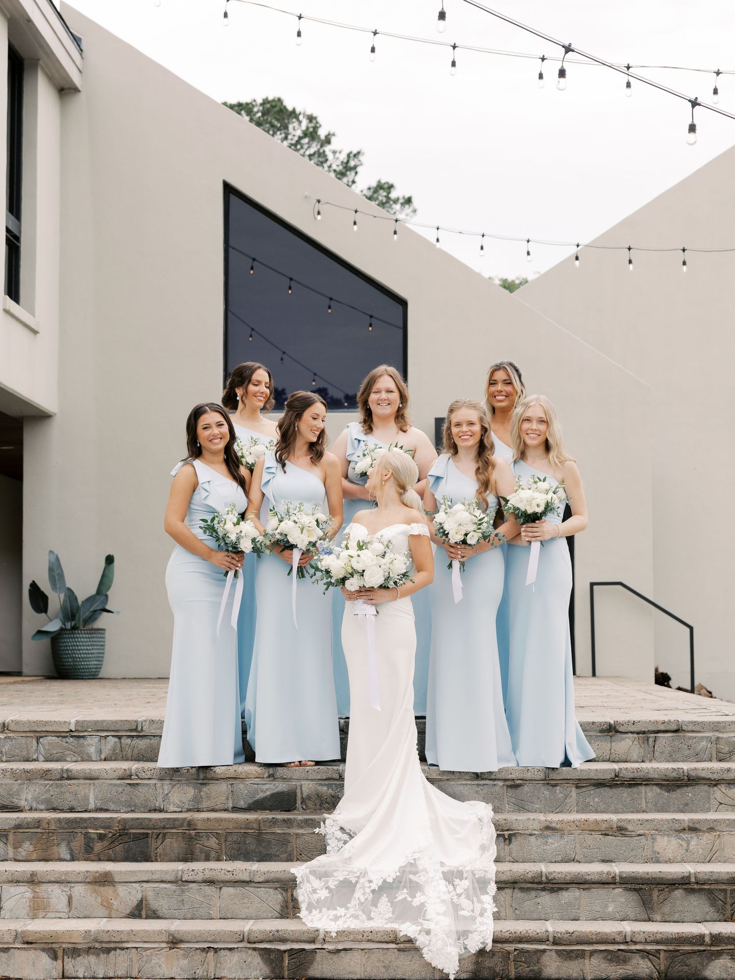 Wedding party in light blue bridesmaid dresses pose on stone steps with string lights overhead.