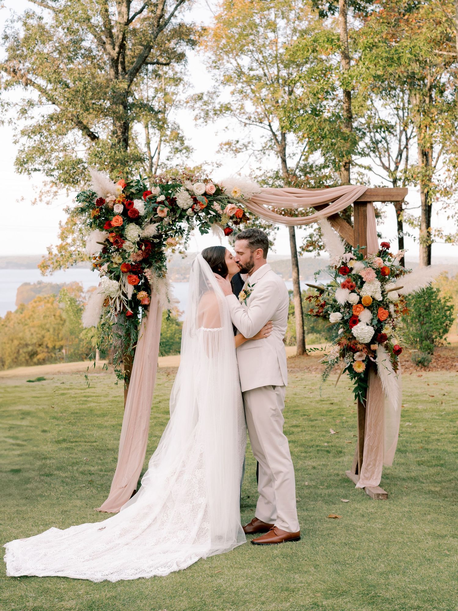 Outdoor wedding ceremony with floral arch and couple exchanging vows on grassy hillside during autumn.