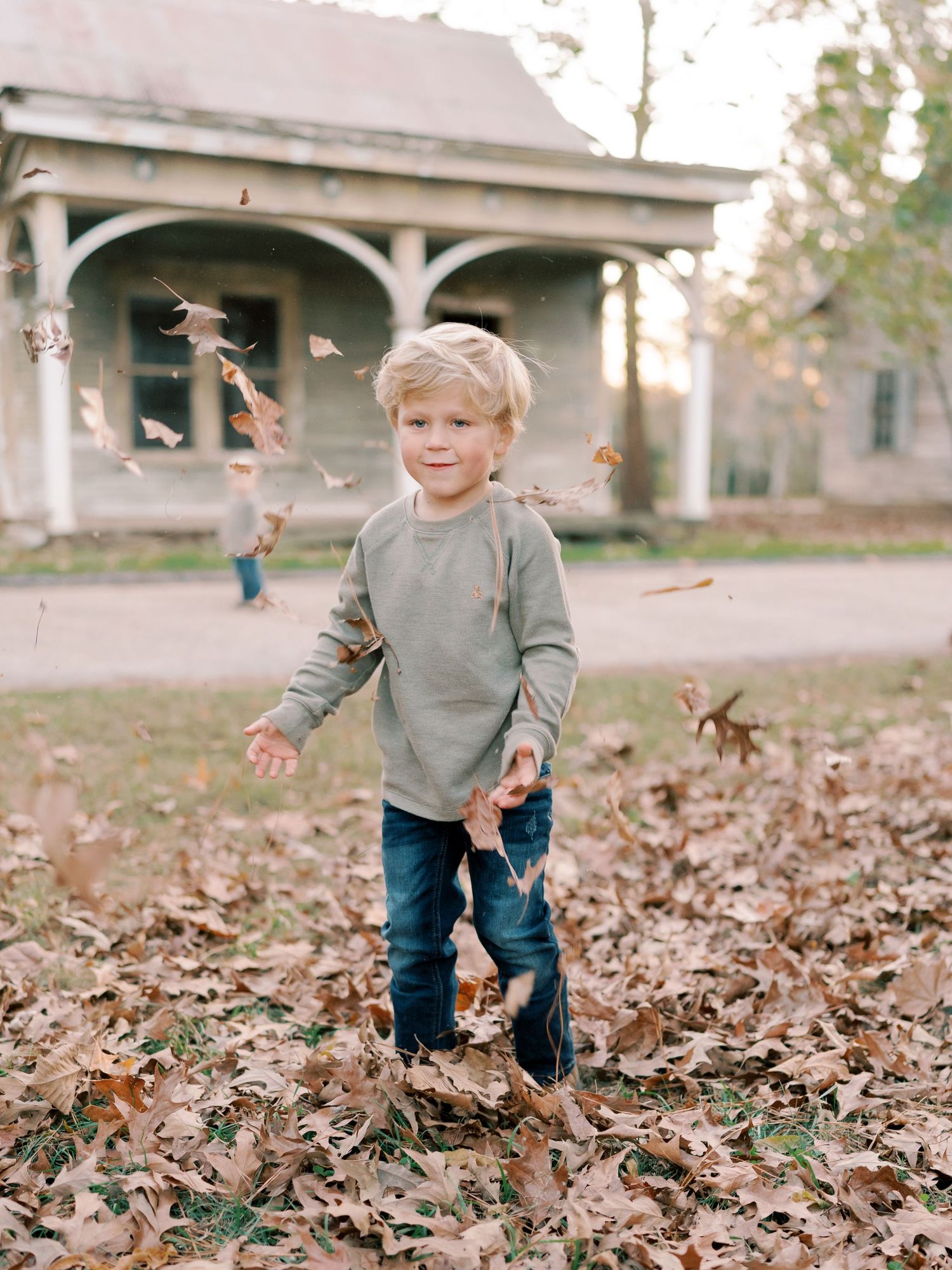 A small child plays joyfully in a pile of autumn leaves near an old house.