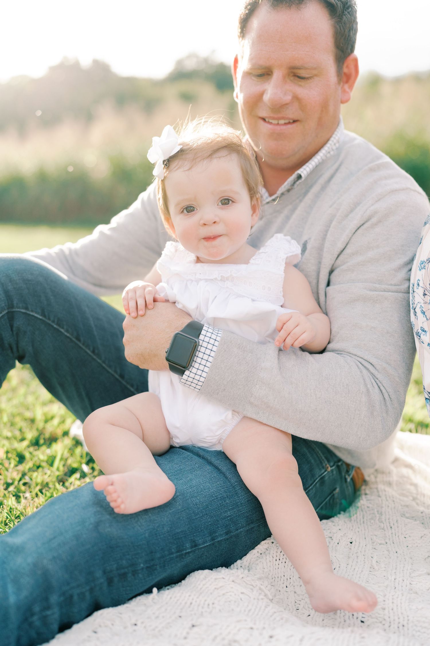 Baby in white dress sits with adult on blanket in grassy field during golden hour.