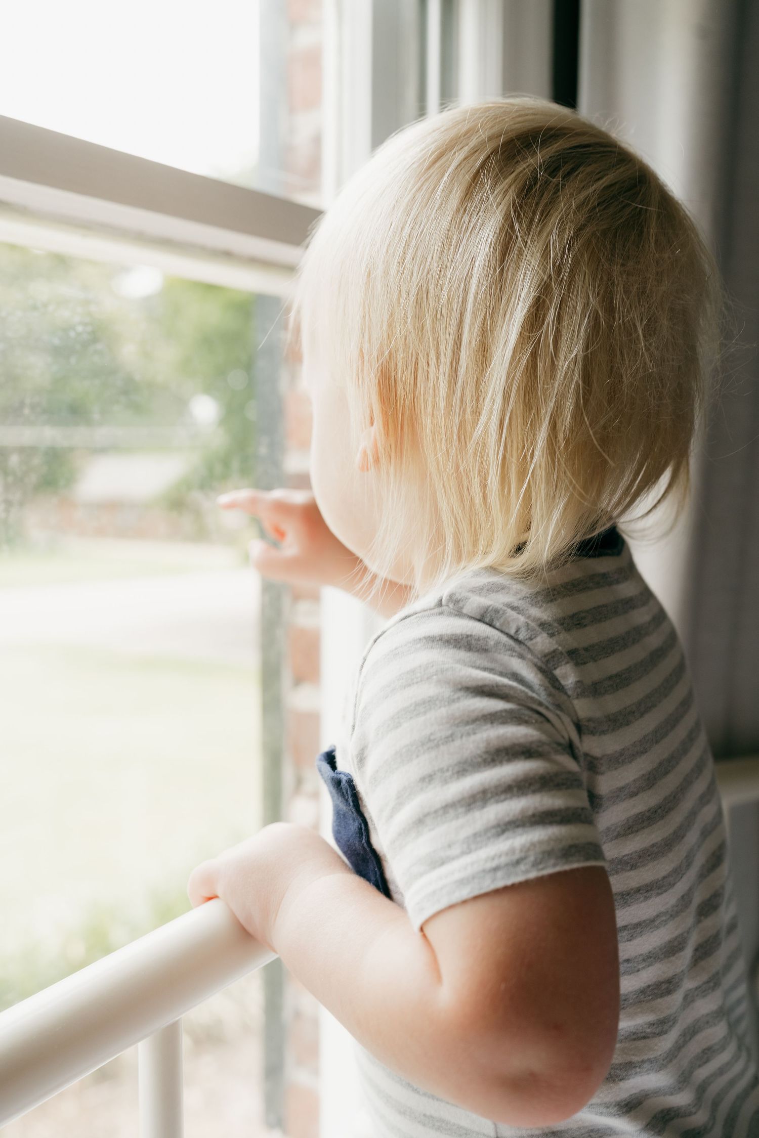 Young child in striped shirt looking out bright window.
