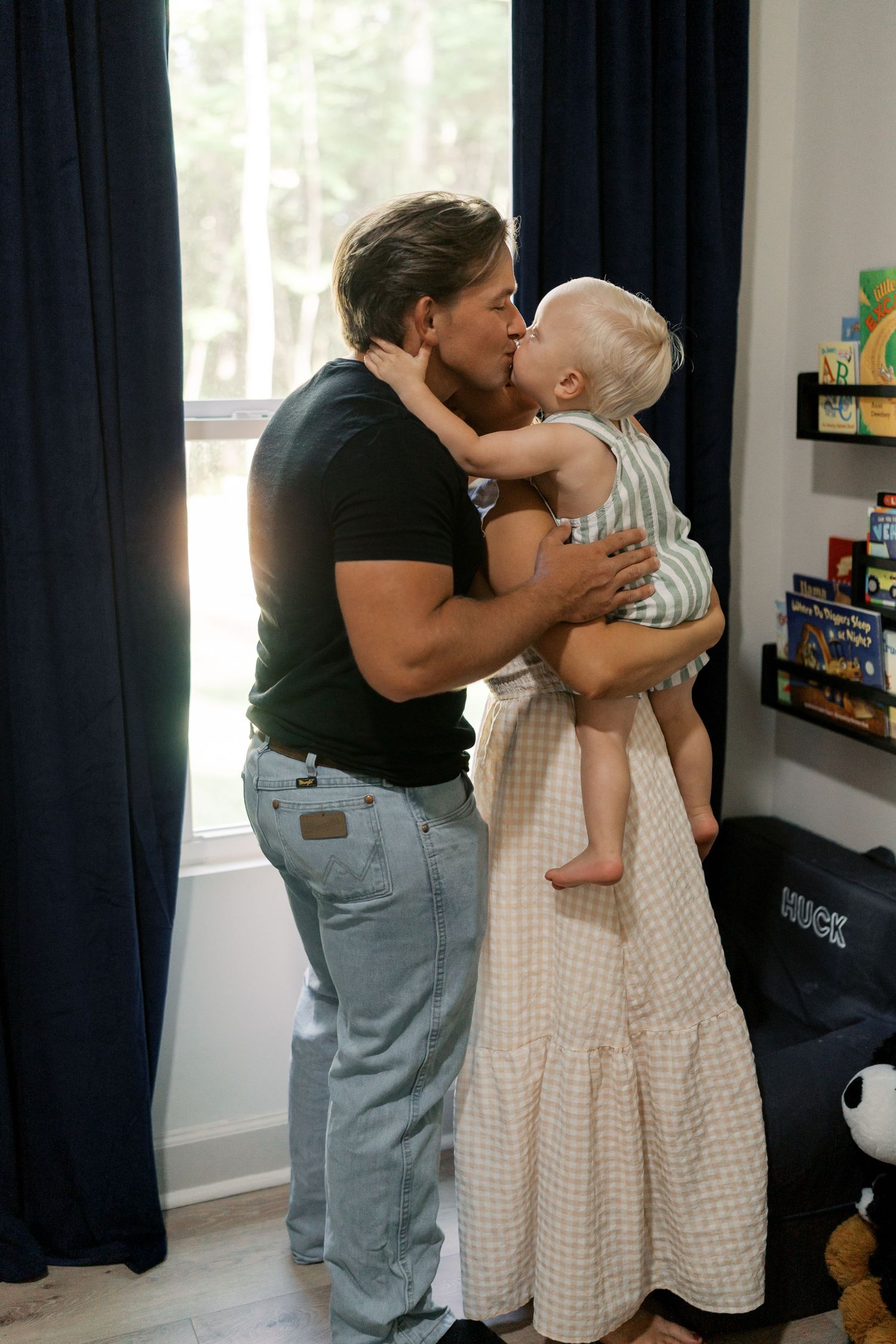 Loving family embraces and kisses in a nursery room with dark curtains and toys on shelves.