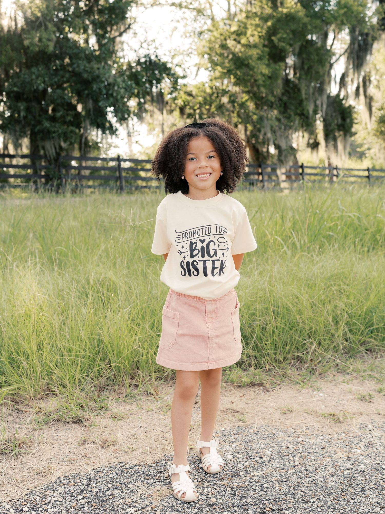 A child wearing a 'Big Sister' t-shirt poses happily on a gravel path outdoors.