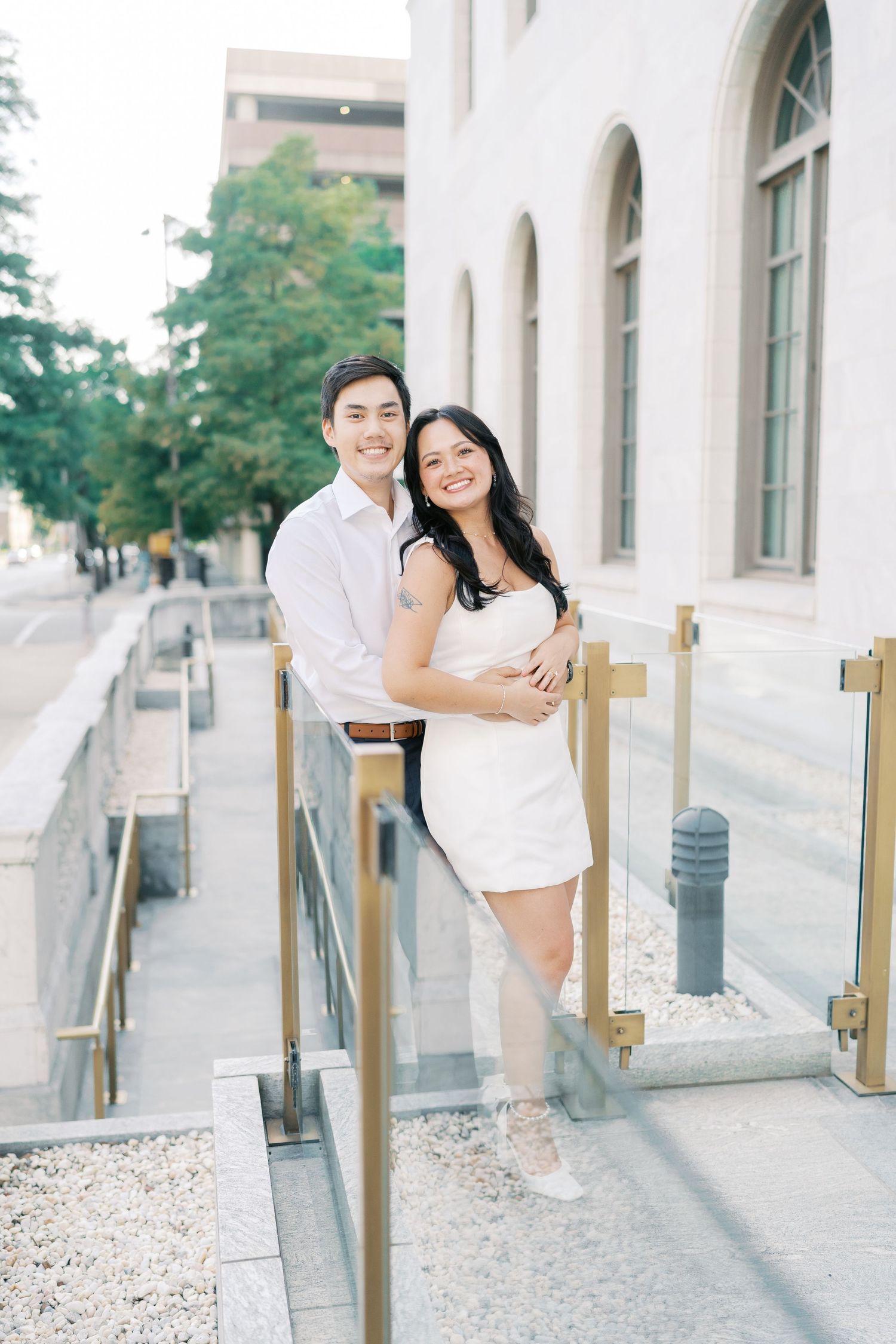 A playful couple poses along a railing outside a classic building with white columns.