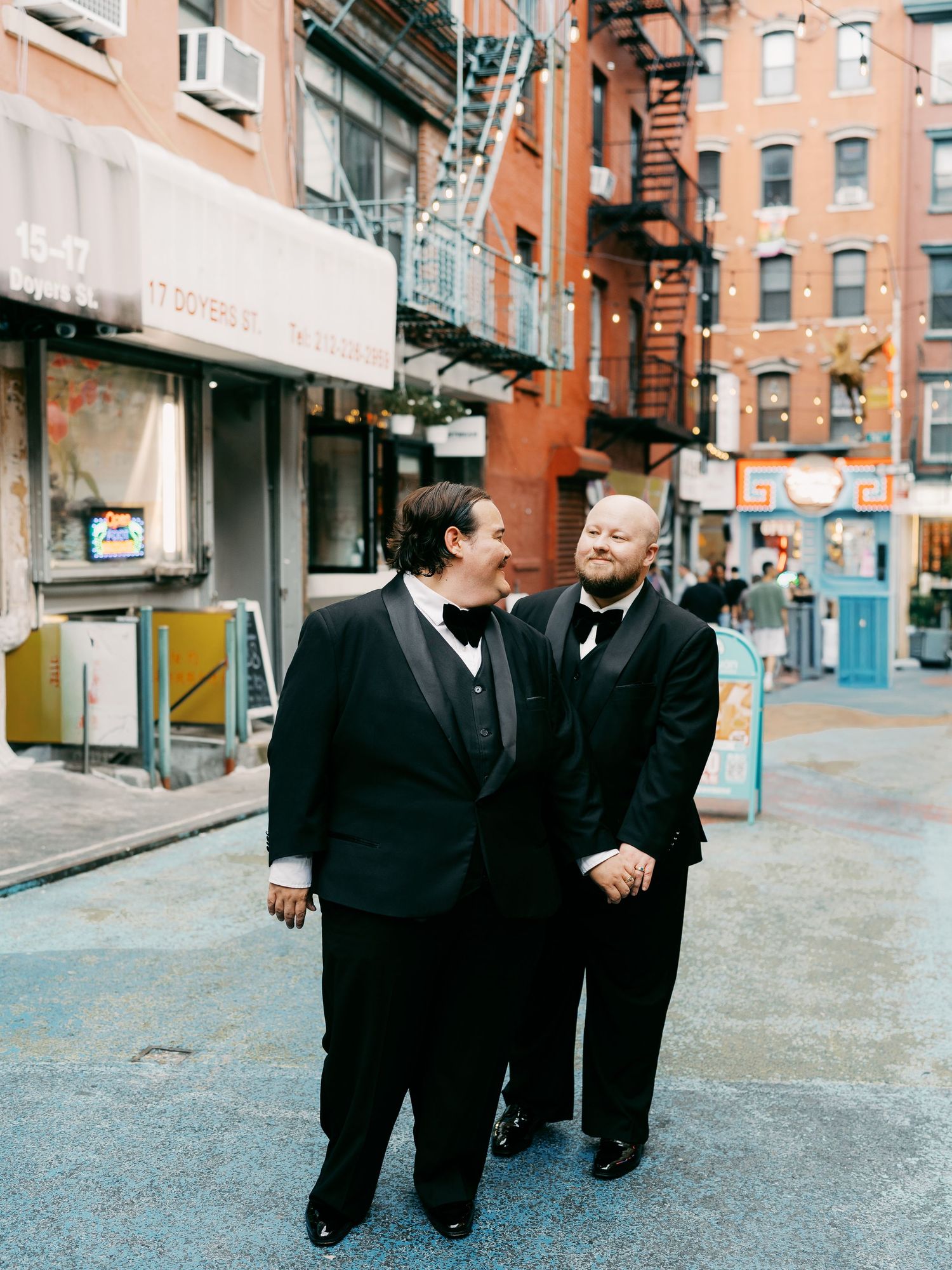 People in dark suits walking along a brick building with fire escapes.