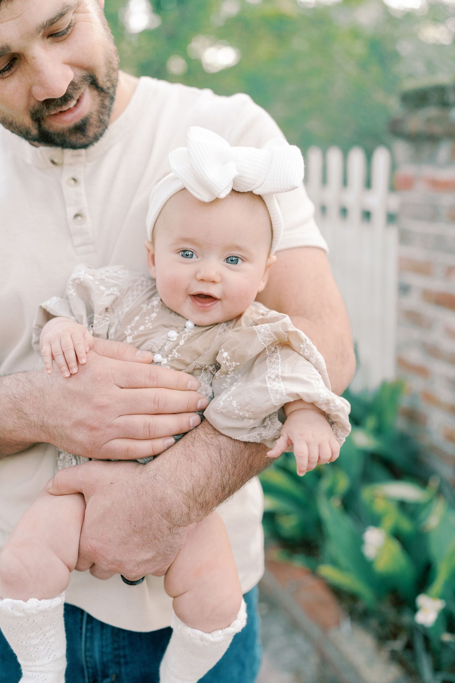A baby in a lace outfit smiles while being held outdoors near a garden.